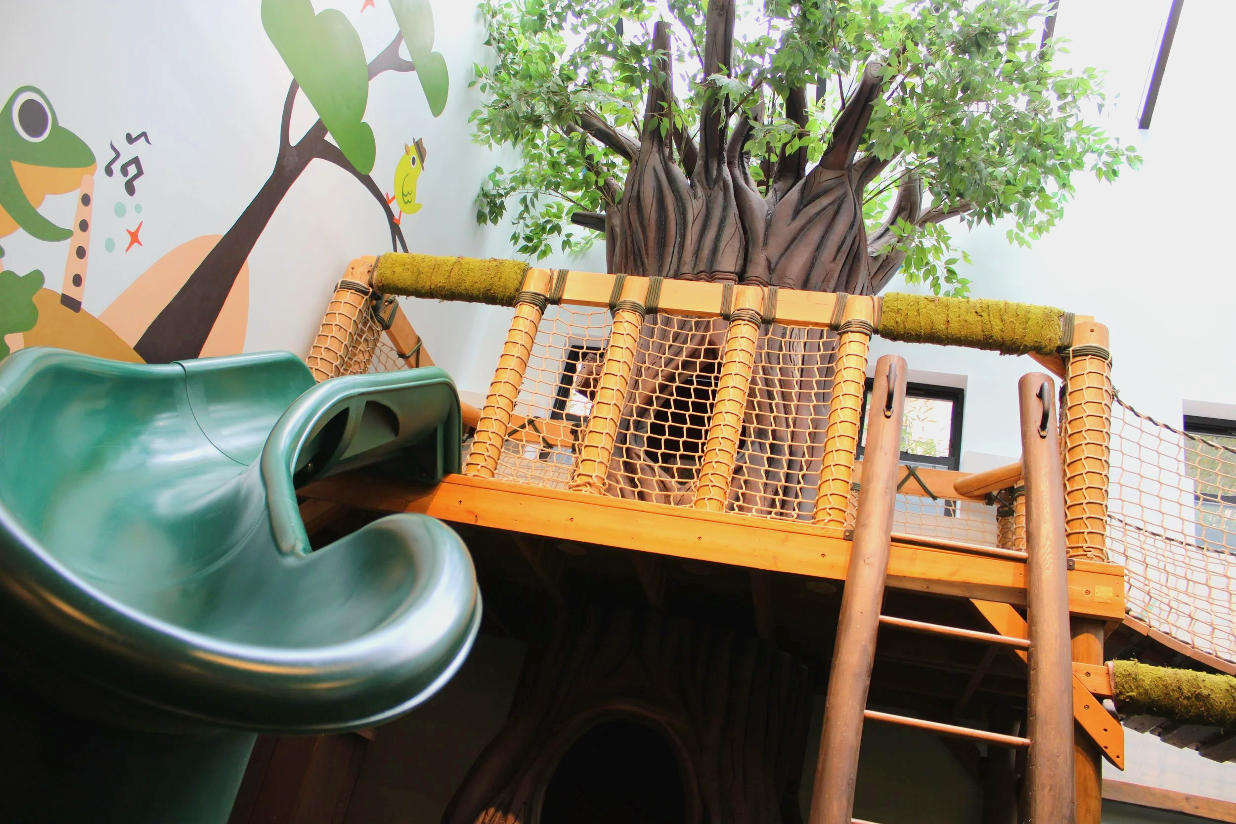 Indoor play area featuring a wooden treehouse structure with a slide, surrounded by painted mural walls with trees and birds.