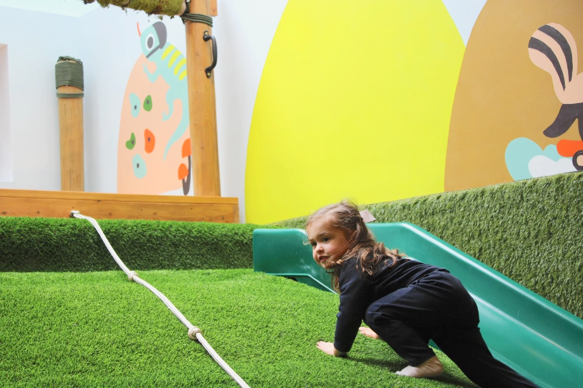 A young girl crawling on artificial grass next to a small green slide, with a background featuring colorful wall art of animals and greenery.