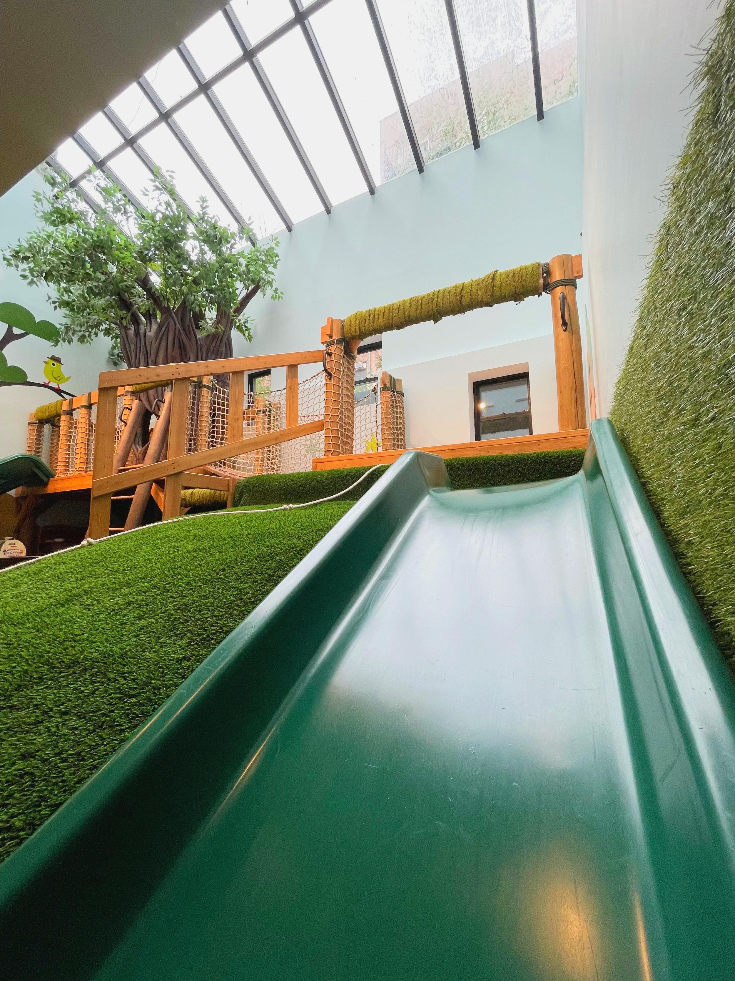Indoor playground slide with a green surface, artificial grass walls, and a wooden treehouse structure with a rope bridge under a glass ceiling.