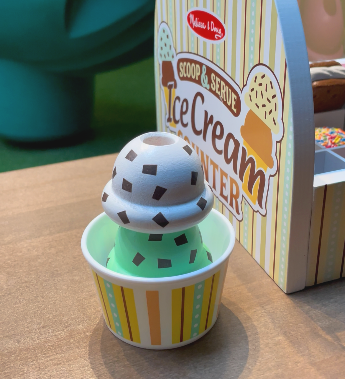Toy ice cream scoop in a striped cup, with a colorful ice cream shop sign in the background.