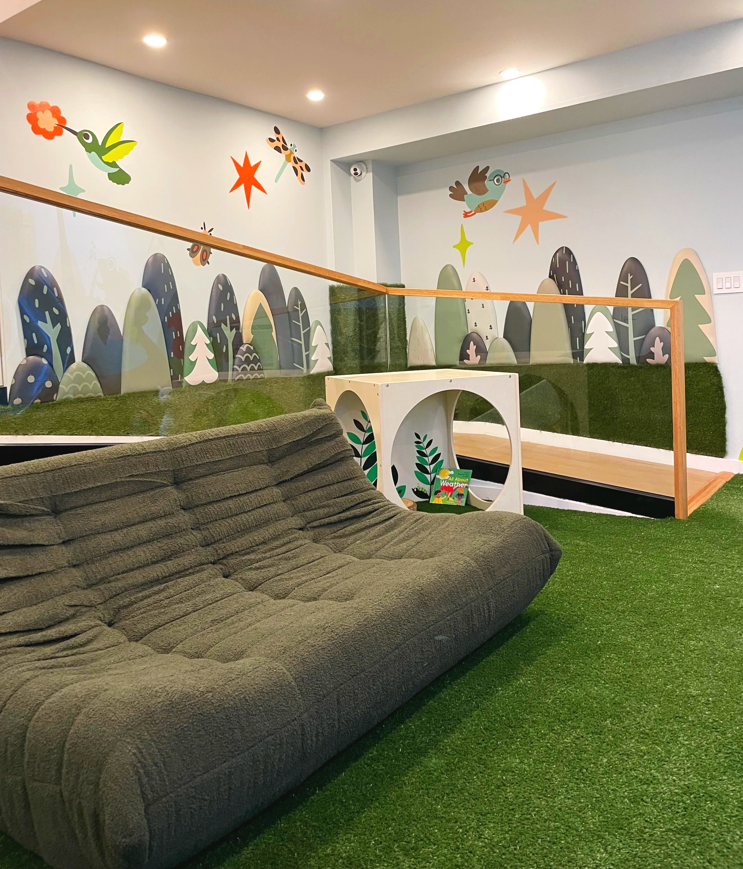 Indoor play area with a mural of stylized trees, mountains, and flying insects, featuring a soft foam climbing structure on green turf.