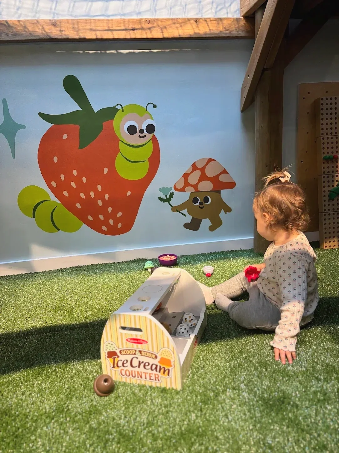 A young girl sitting on artificial grass with a toy ice cream counter, looking at a colorful mural of cartoon bugs and strawberries on a wall.
