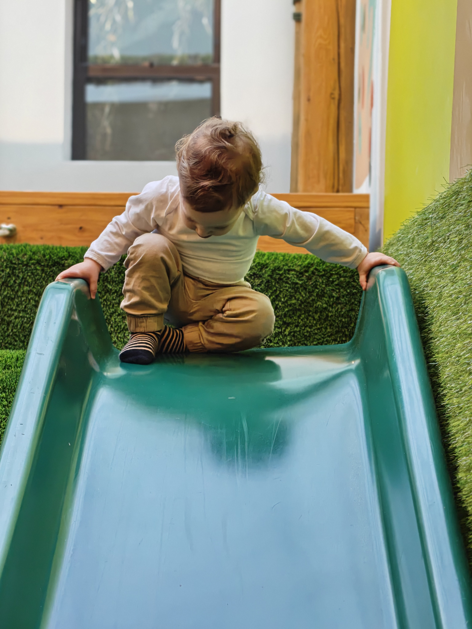 A young boy with light brown hair wearing a white long-sleeve shirt and tan pants crawling up a small green plastic slide in an indoor play area with green artificial grass flooring.