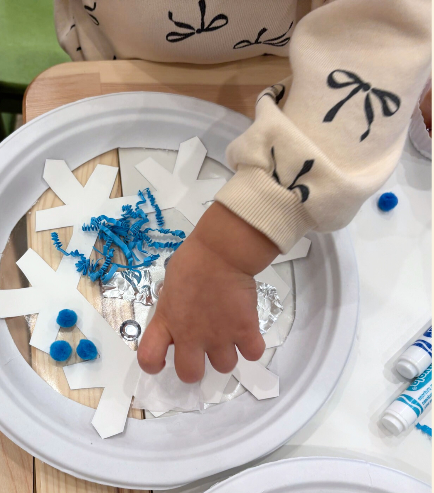 Child wearing a beige sweatshirt with black bow designs, reaching into a cutout of a snowflake-shaped paper craft with blue tissue paper and pom-poms inside, on a wooden table.