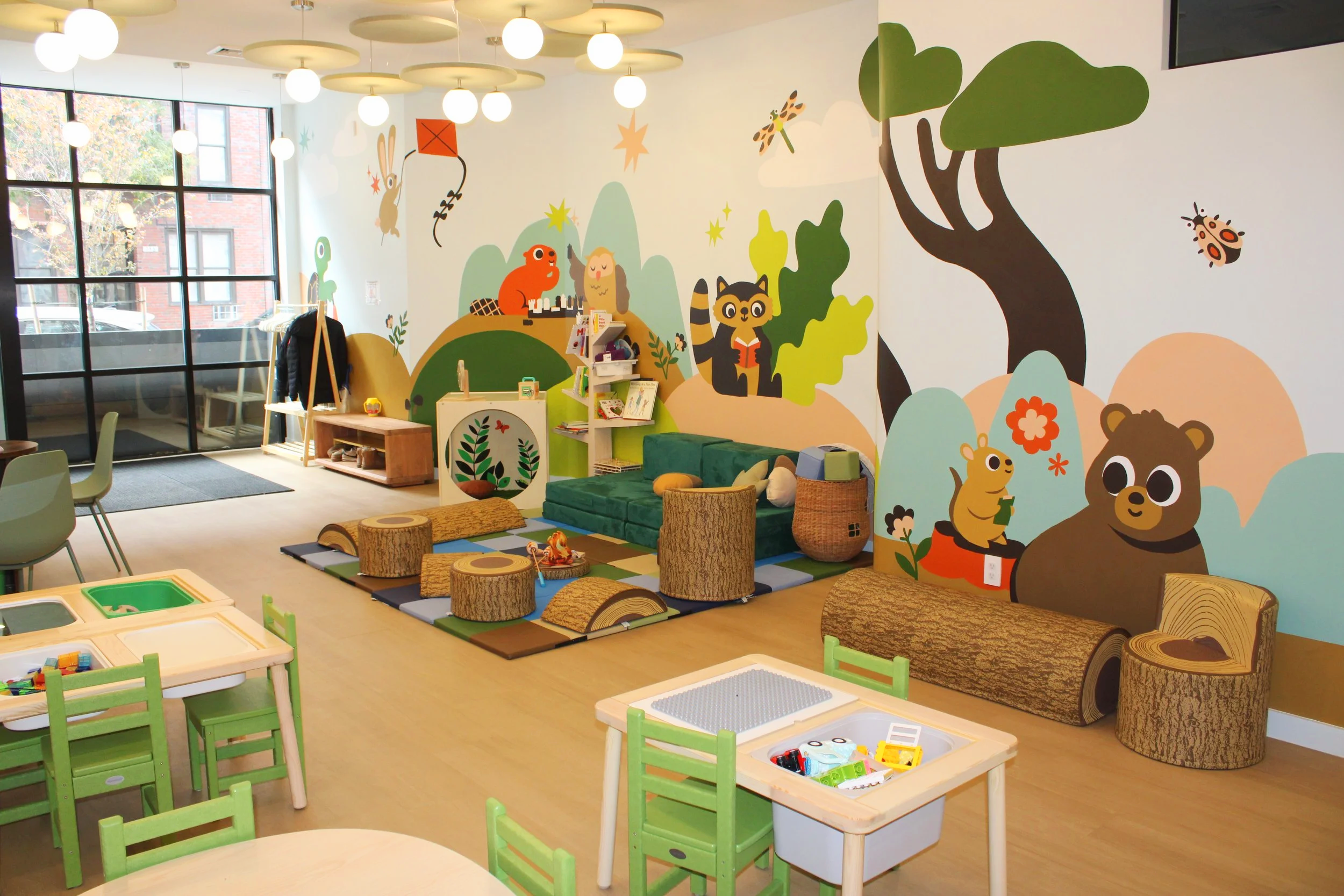 A children's playroom with colorful wall murals of animals, trees, and nature scenes, featuring a green couch, small tables, chairs, and a variety of toys and books.