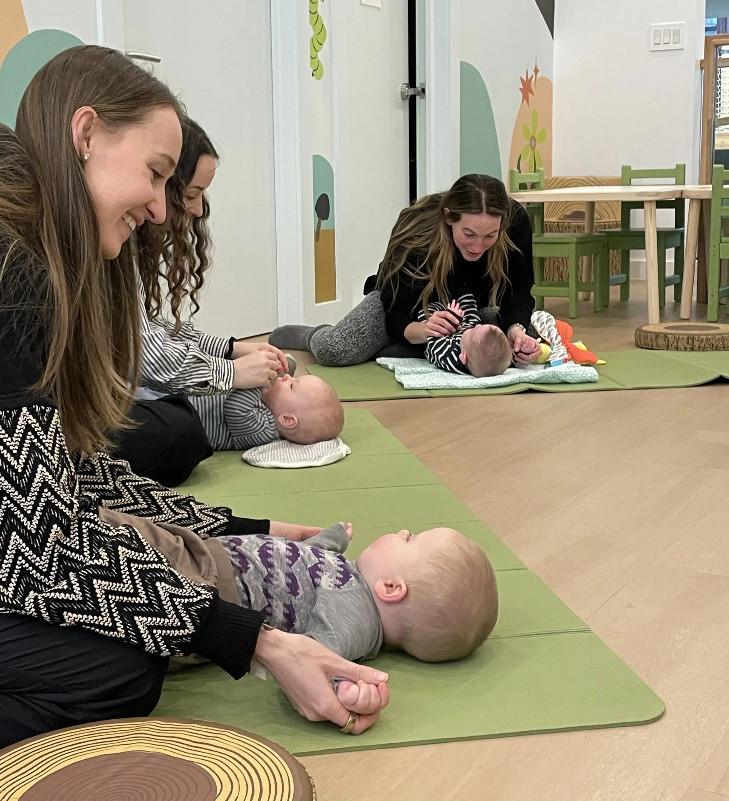 Women and infants engaging in a practice session on yoga mats in a daycare or playroom setting, with playful wall art and furniture in the background.