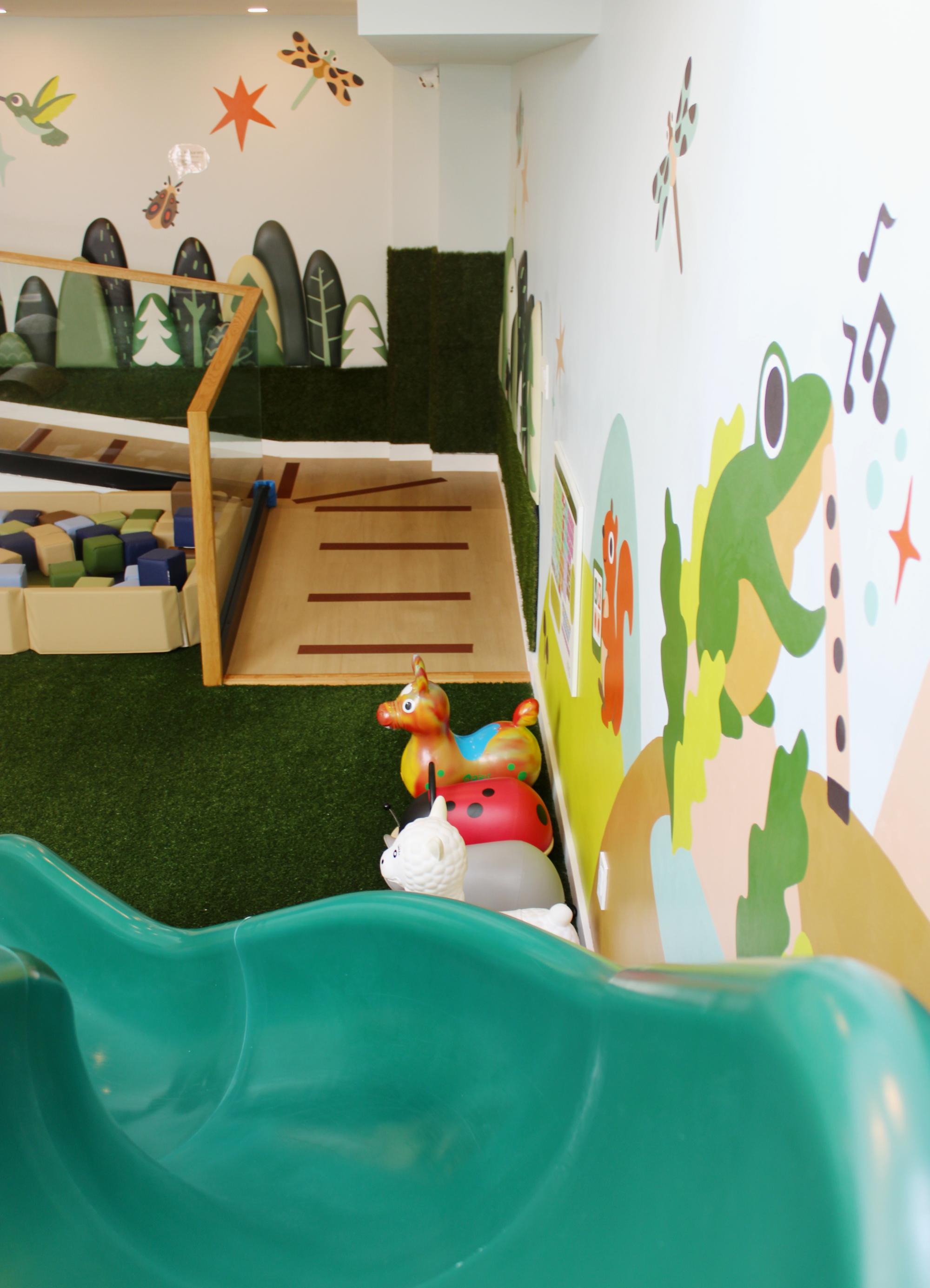 Interior of a children's playroom with a green slide at the bottom, colorful animal and nature-themed wall art, and inflatable animal toys