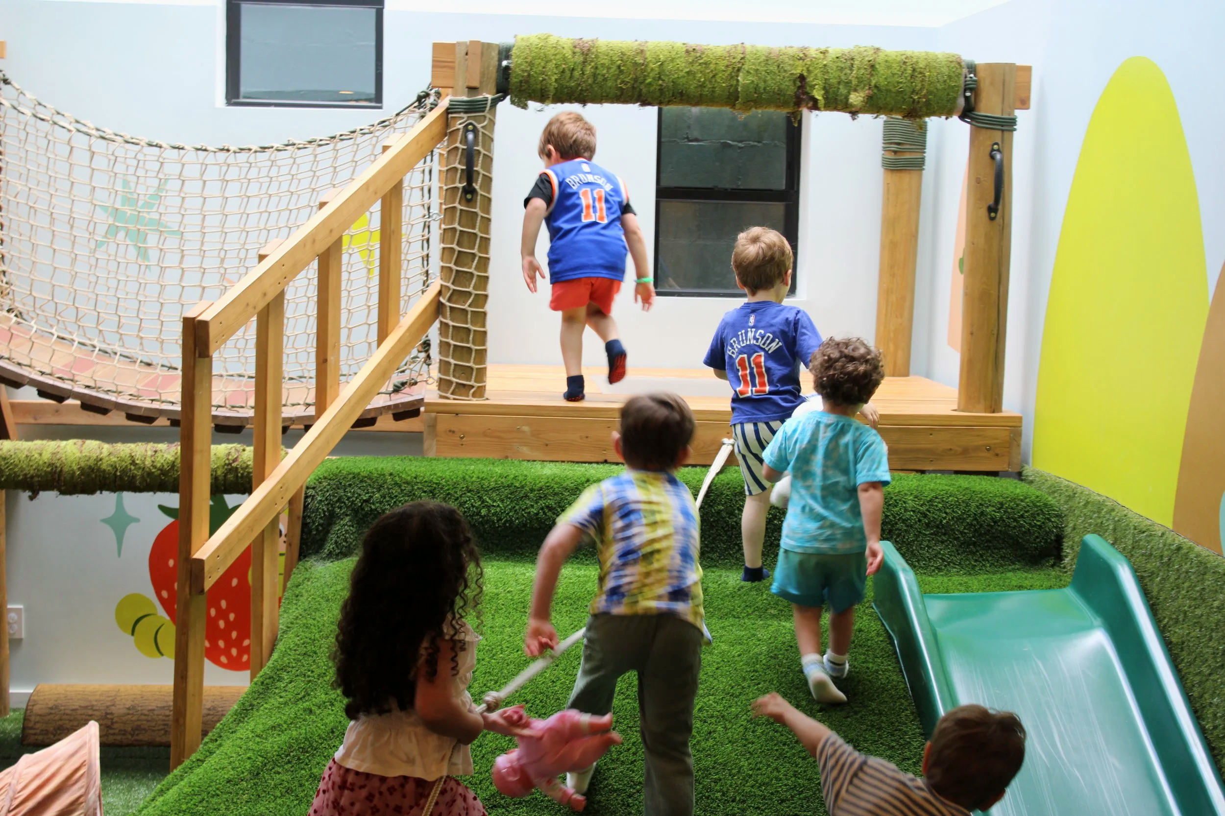 Children playing at an indoor playground with wooden climbing structures, a green slide, and artificial grass flooring.