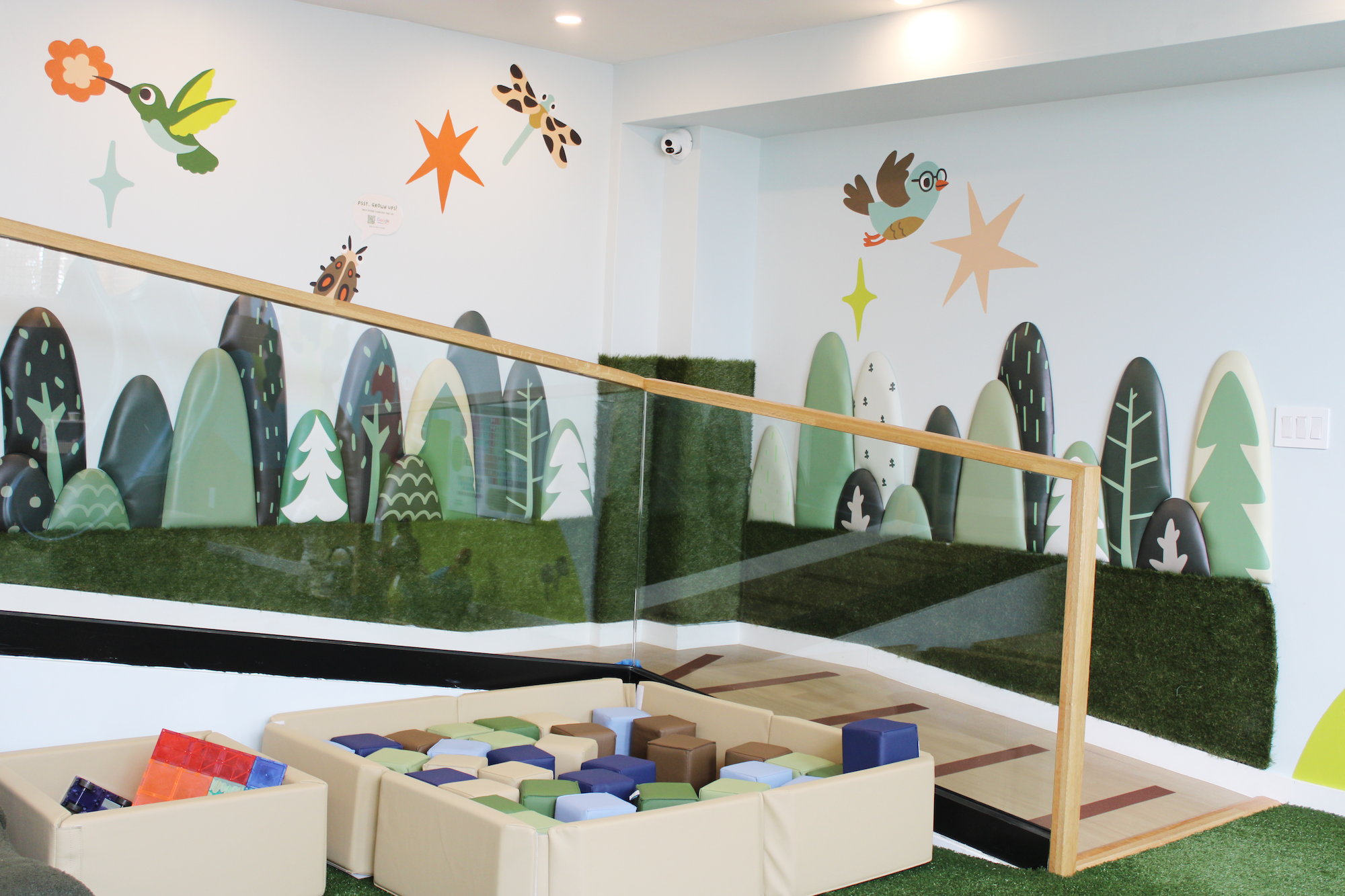 Colorful indoor children's play area with woodland themed wall art, including owls, hummingbirds, stars, and trees, with a padded area for play and wooden railings.