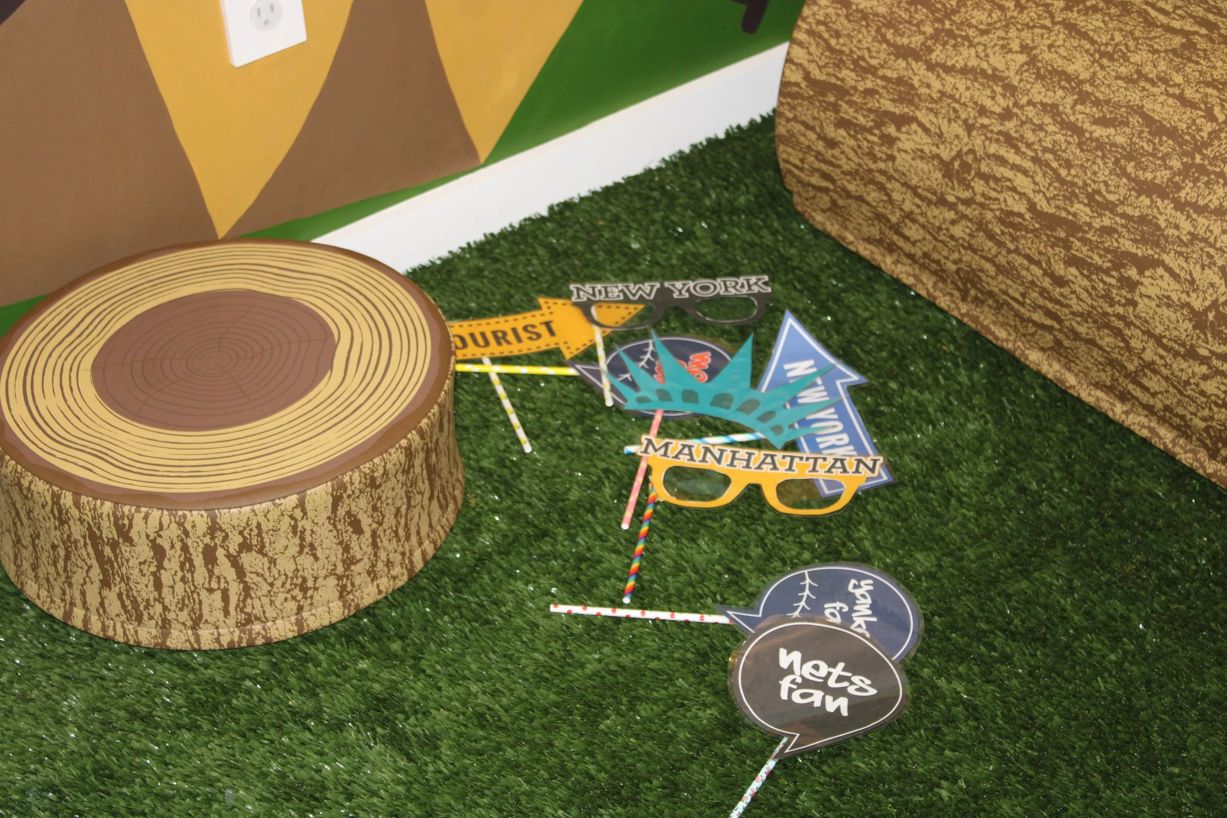 Decorative photo booth backdrop with artificial grass floor, wood tree stump prop, and colorful paper props including glasses with 'New York,' 'Manhattan,' and 'Tourist' labels, and speech bubble signs with congratulatory messages.