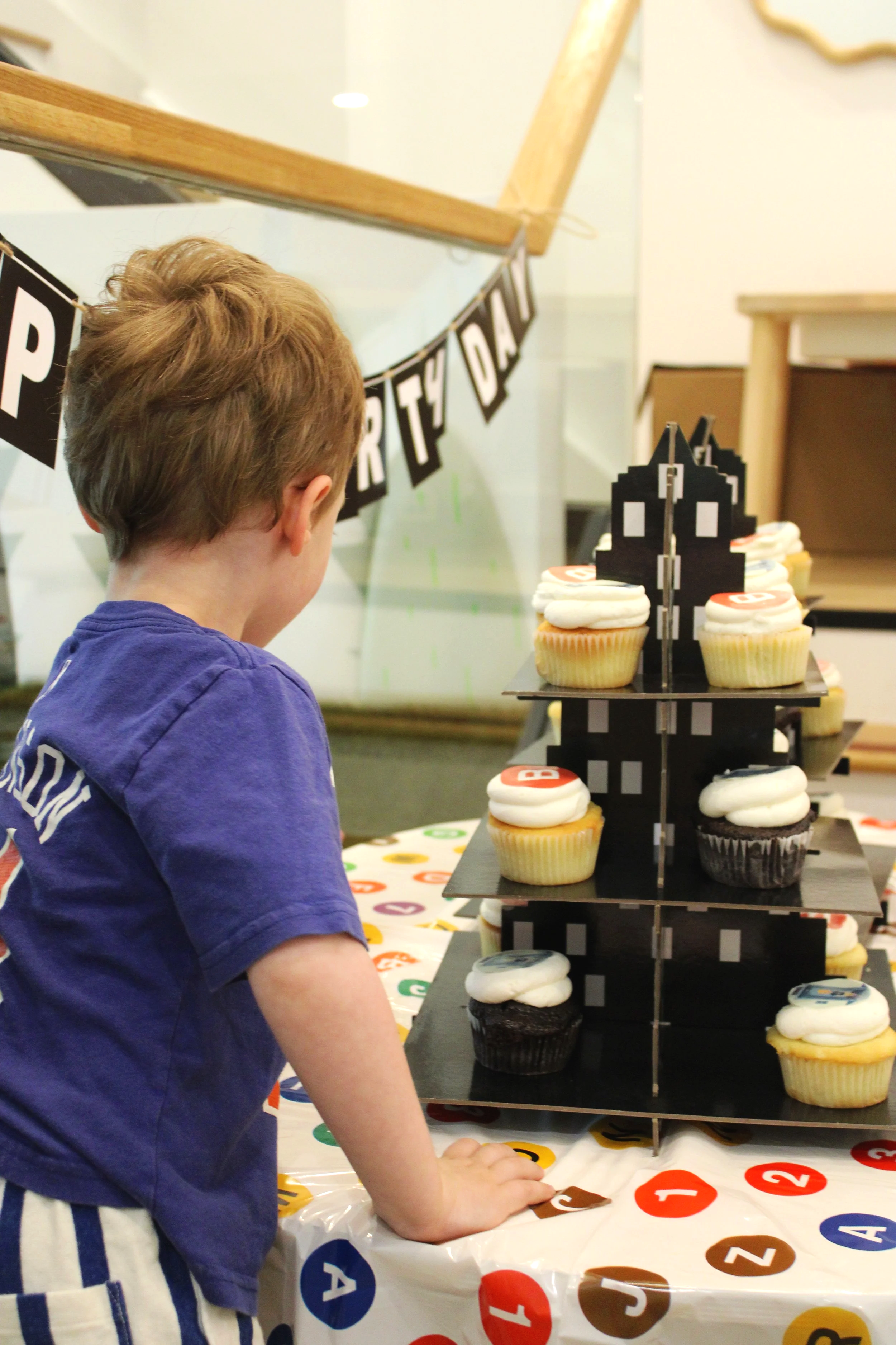 A young boy wearing a blue shirt the cupcake stand has black tiers with cupcakes decorated with white or dark chocolate frosting. A banner with black squares and white letters spelling "Happy Birthday" is hung in the background.
