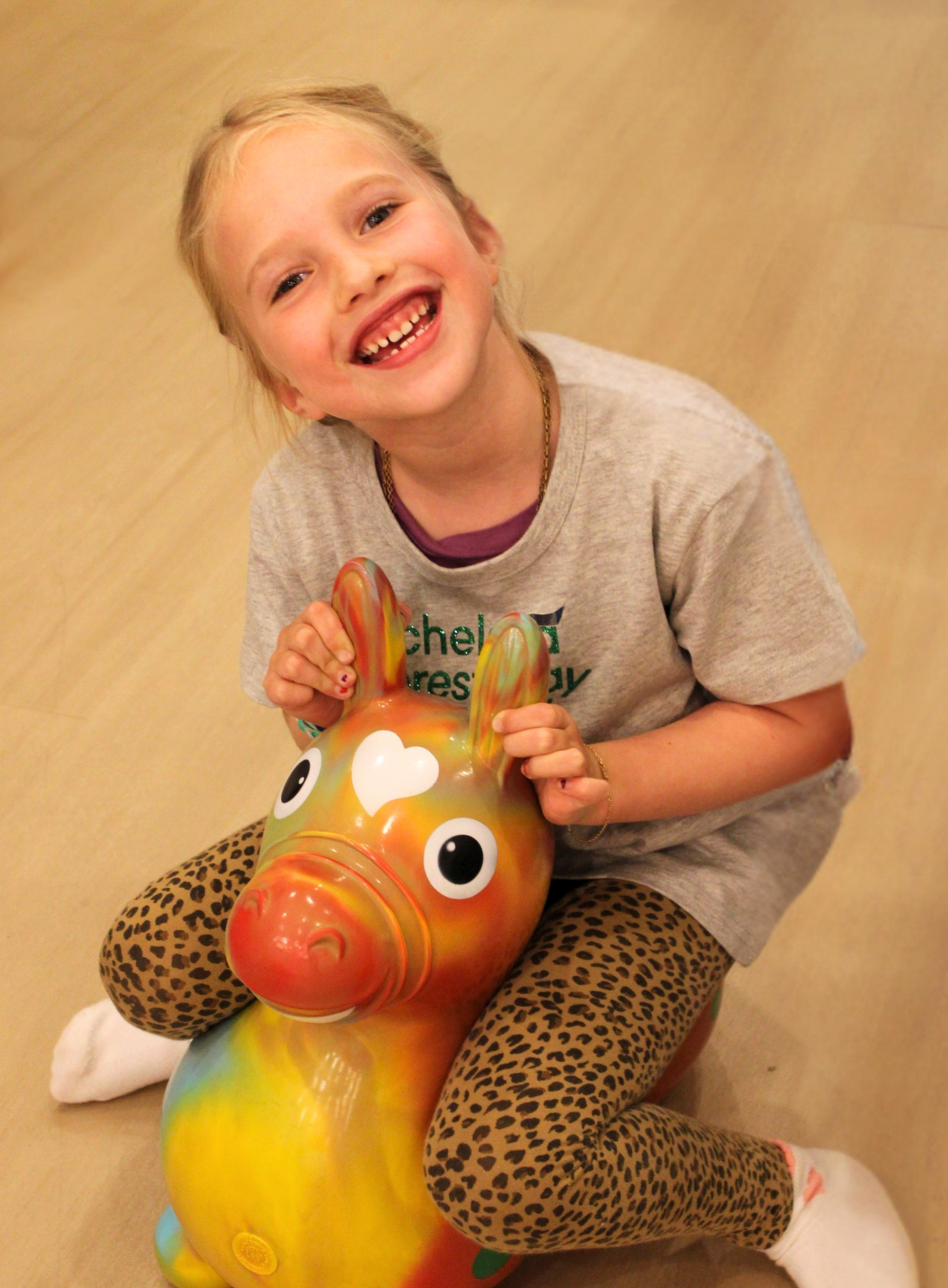 A smiling young girl with blonde hair holding a colorful animal-shaped balloon, wearing animal print pants and a gray T-shirt, sitting on a beige floor with a plain beige background.