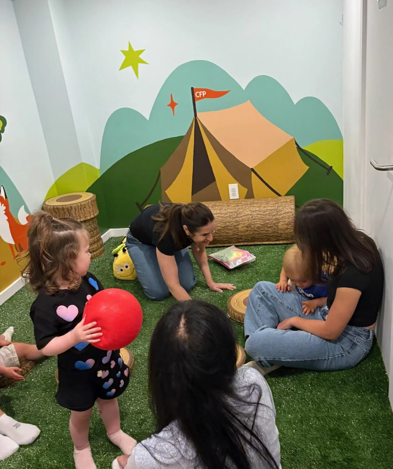 Children and adults sitting on artificial grass in a room with mural walls depicting mountains, stars, and a tent, engaging in a group activity.