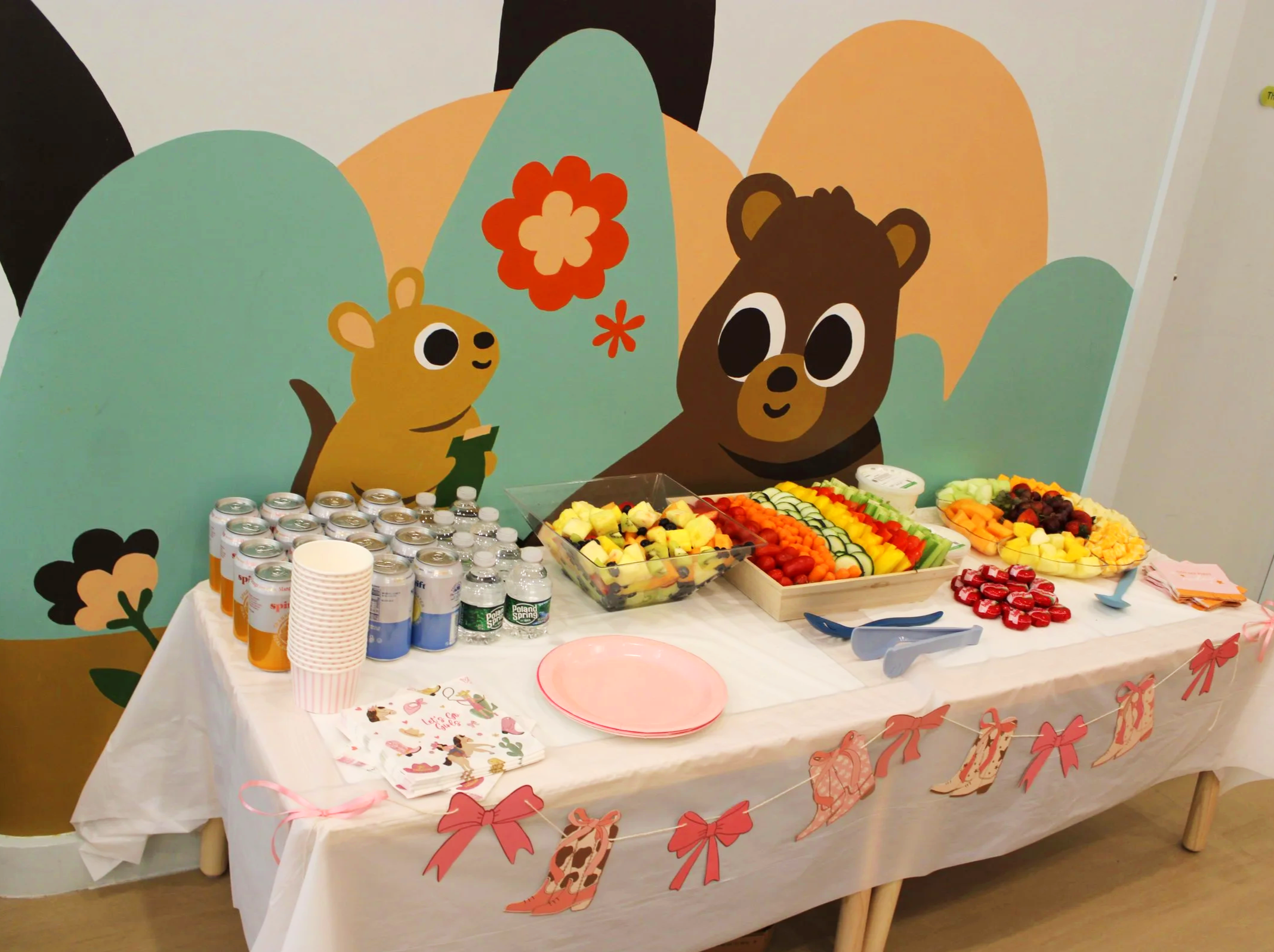 A table decorated with pink bows and a bunting, holding a variety of food including cut fruits, small desserts, and cans of soda, set against a colorful mural featuring cartoon animals and plants.