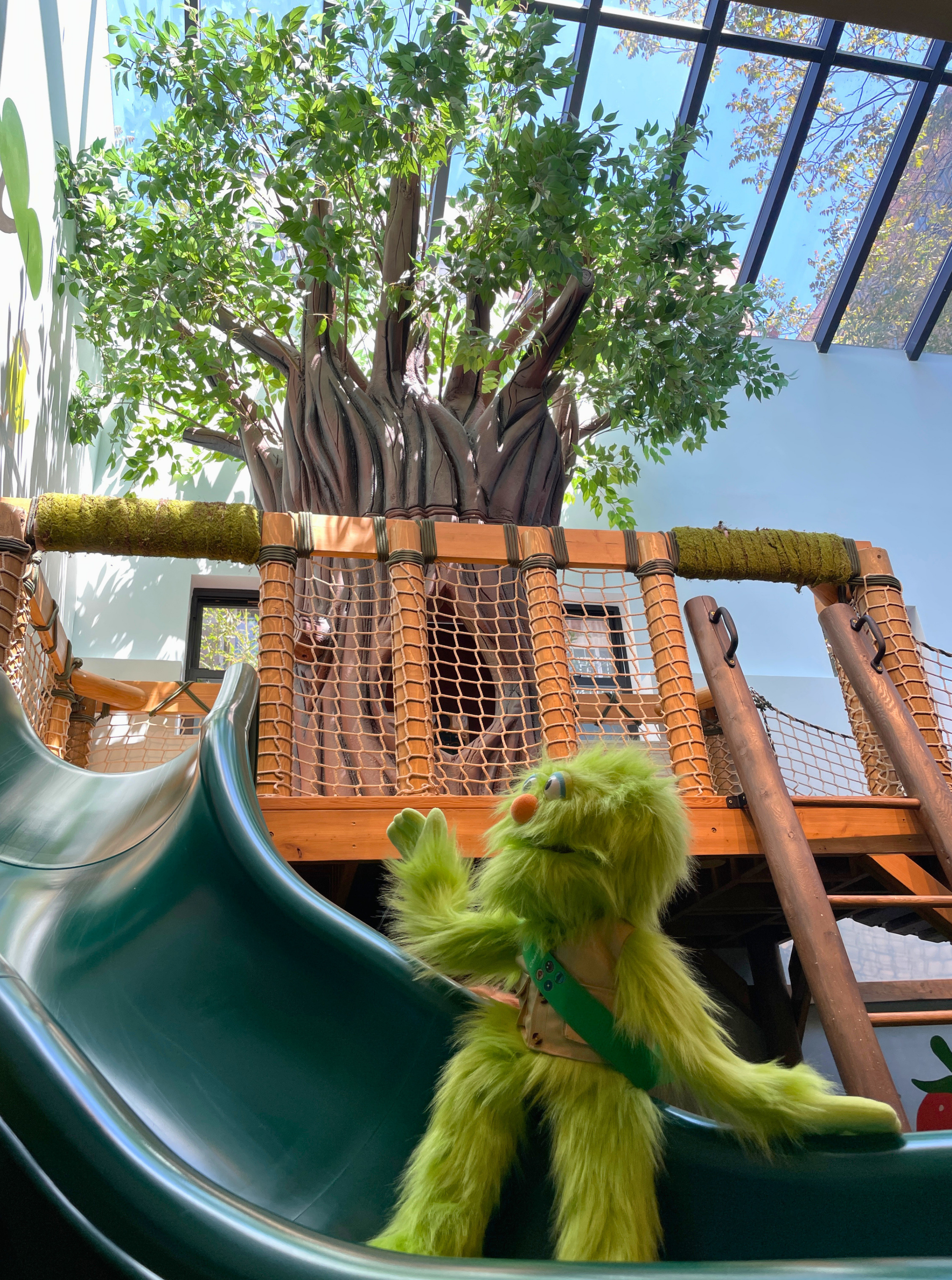 A green furry puppet with large eyes and an orange nose, sitting at the bottom of a playground slide, with a large indoor tree and glass ceiling in the background.