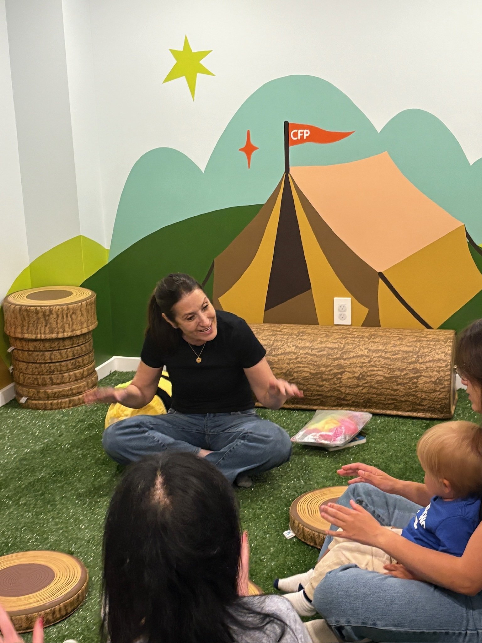 A woman with dark hair sitting on the floor surrounded by children, with a colorful mural of mountains, stars, and a tent in the background.