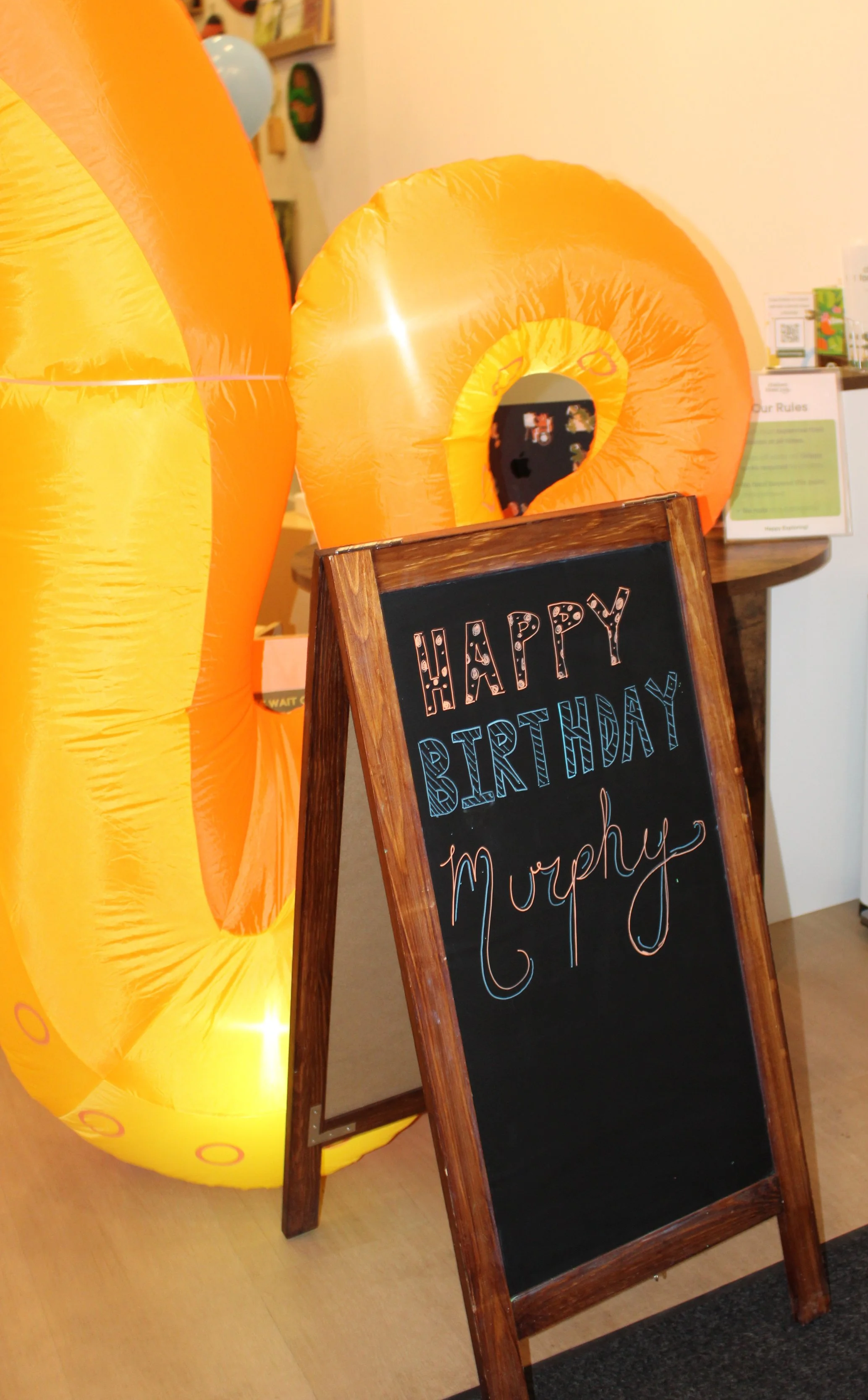 Decorative inflatable banana with a black chalkboard sign that says 'Happy Birthday Murphy' in multicolored writing, set indoors for a birthday celebration.