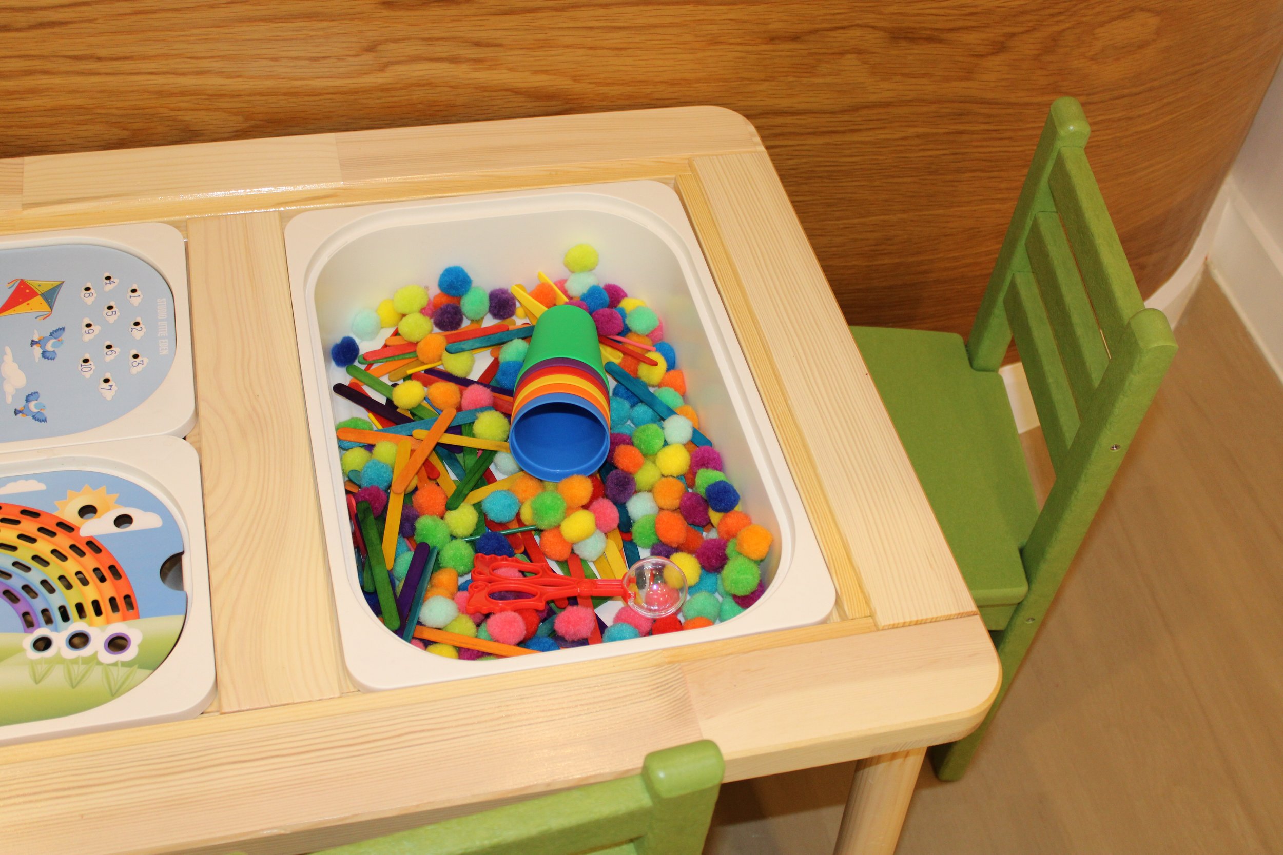 A children's table with a toy box filled with colorful pom-poms and craft sticks, next to a green wooden chair.