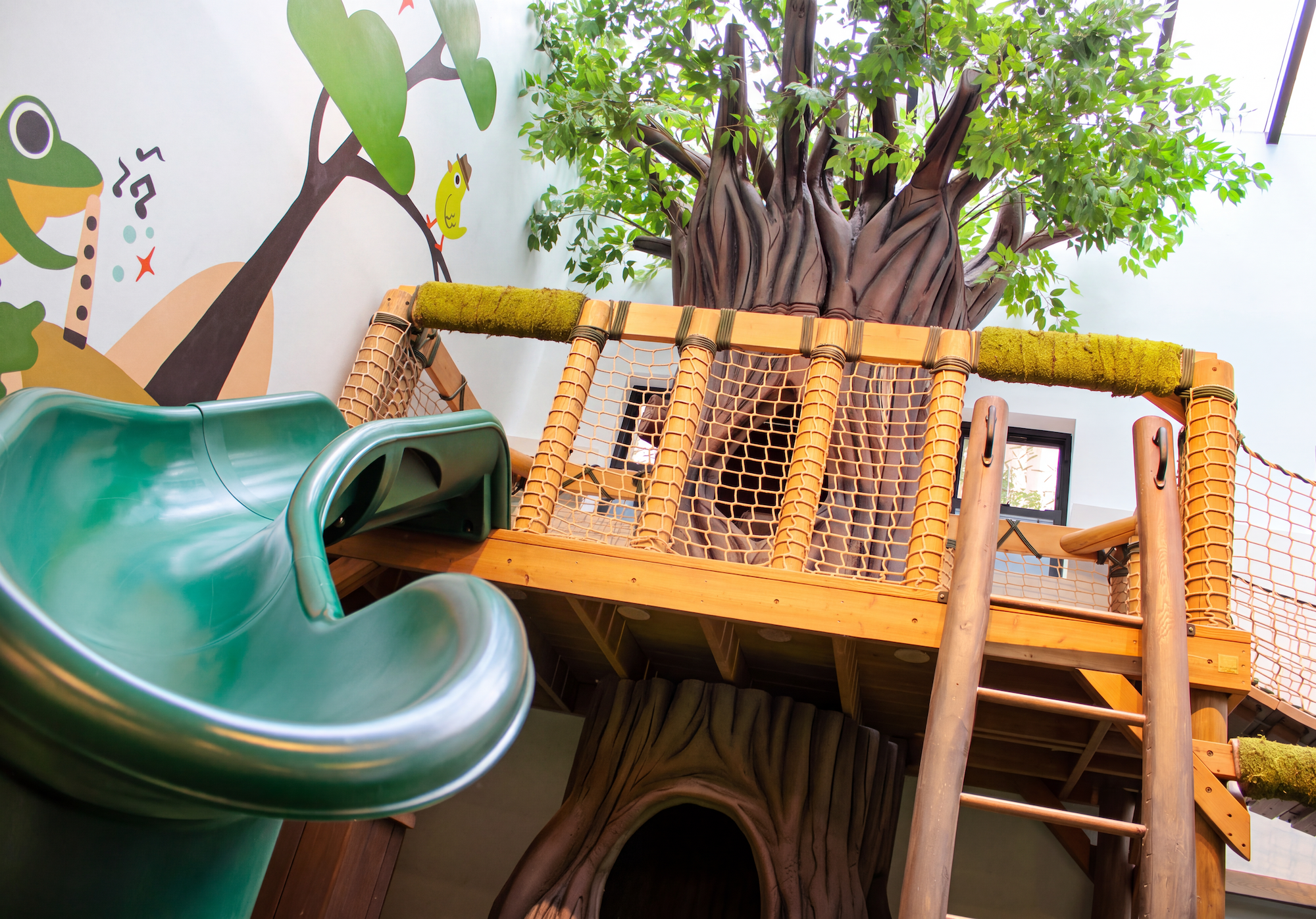 Indoor playground featuring a large tree structure with green leaves, wooden branches, and climbing nets. A green slide is visible in the foreground, and the background includes a painted wall with animal illustrations, including a frog, zebra, kangaroo, and bird.
