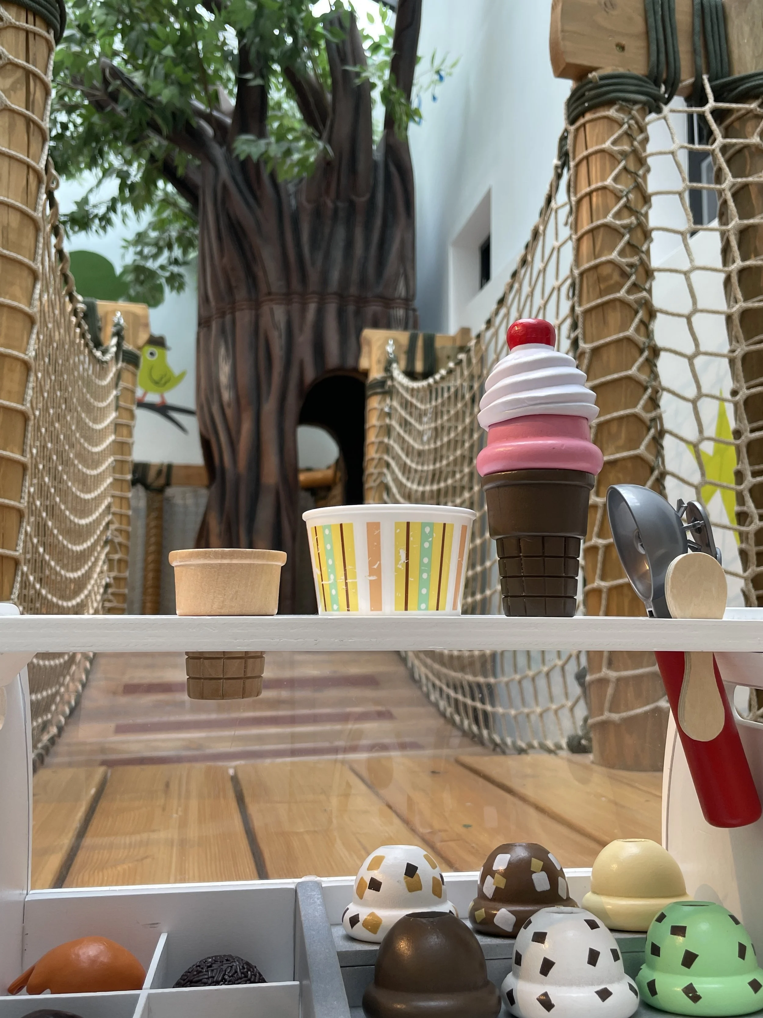 A small display of toy kitchen objects, including a plastic ice cream cone, a paper cup, and several small balls with black and gold patterns, on a white table. In the background, a wooden railing, a wooden floor, and a decorative tree structure.