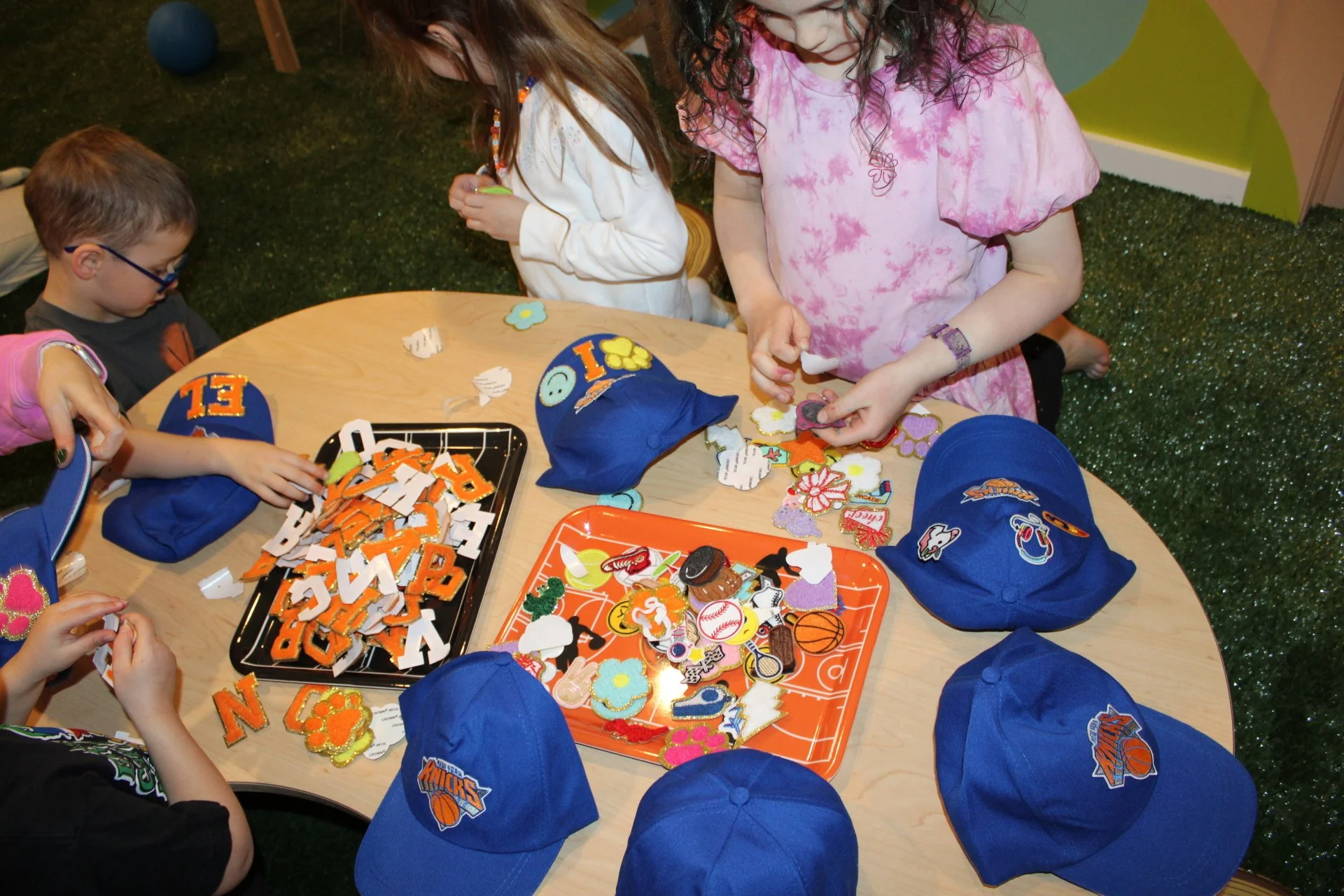 Children gathered around a table decorating with sports-themed patches, hats, and stickers, wearing New York Knicks caps.