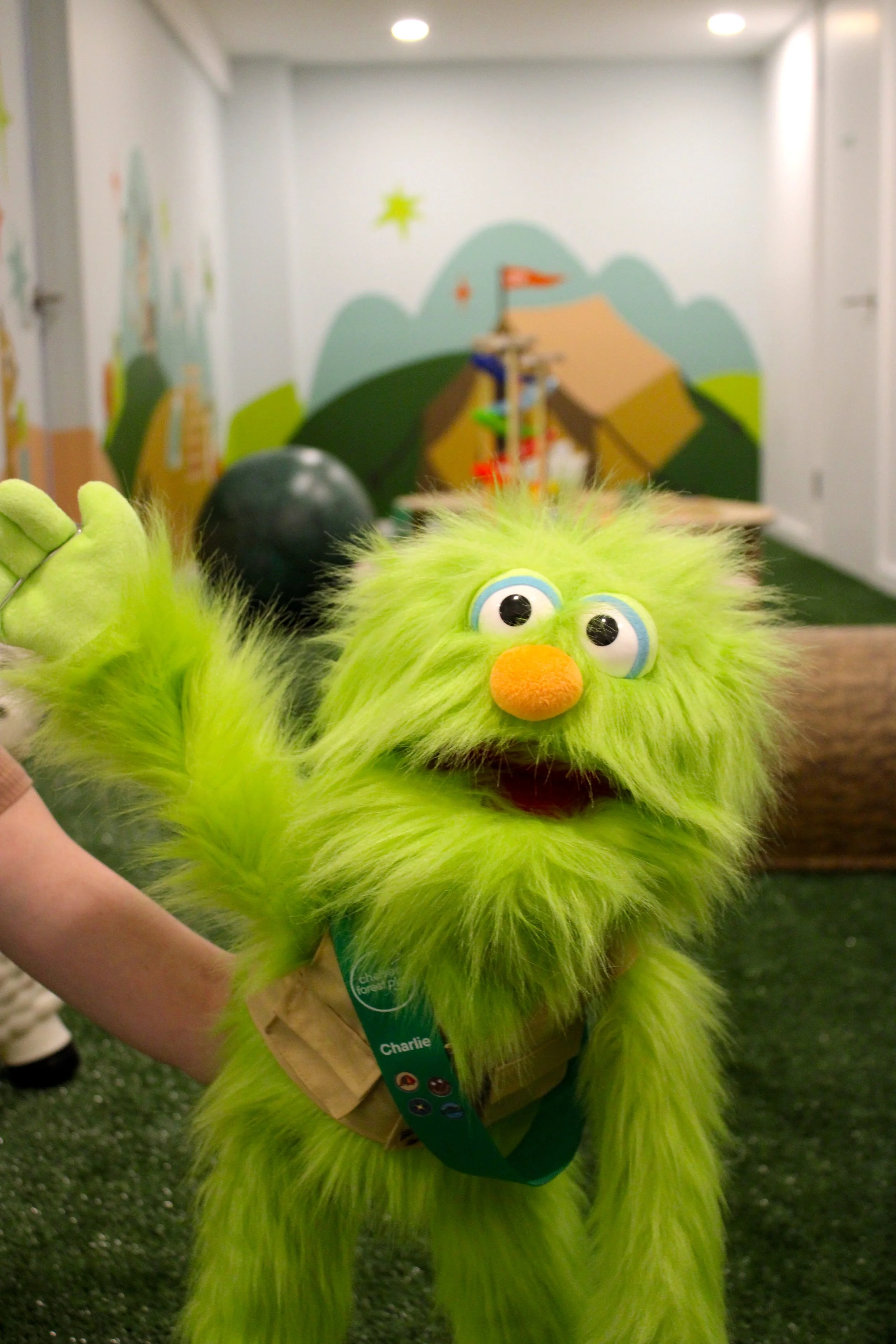 A bright green, furry puppet with large googly eyes, an orange nose, and a big smile, held up by a person wearing a beige outfit. The puppet has a label around its neck reading "Charlie." The background features a colorful mural of mountains, tents, 