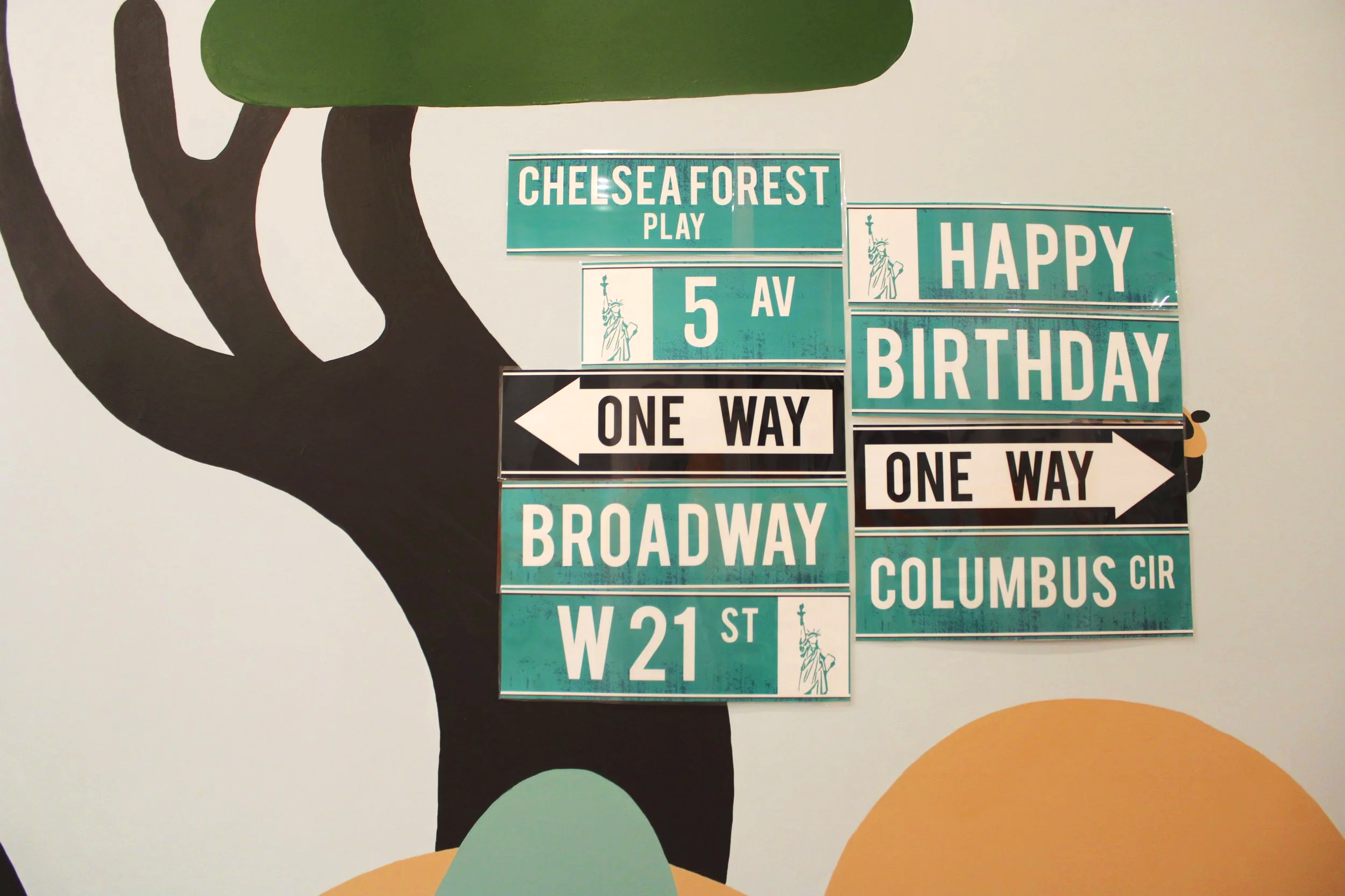A wall decorated with a mural of a woman’s head silhouette and multi-colored circles, with traffic signs and street names including Chelsea Forest Play, 5th Avenue, Happy Birthday, Broadway, and Columbus Circle.