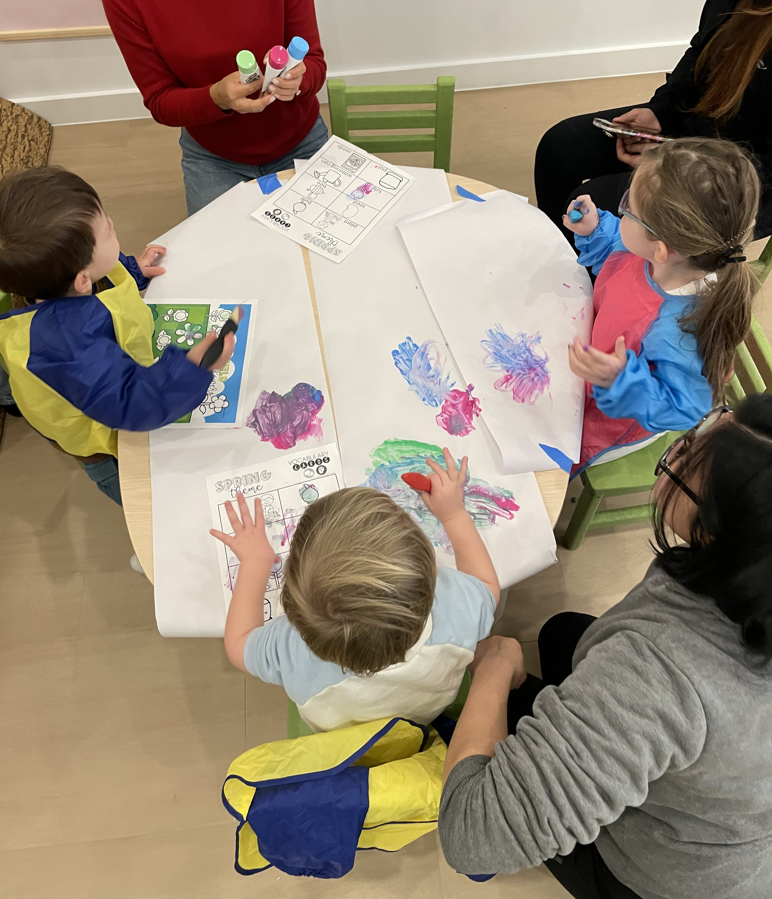 Children and adults sitting around a table with drawings and colorful handprints, engaging in a painting activity at an art class for kids in NYC, at chelsea forest play