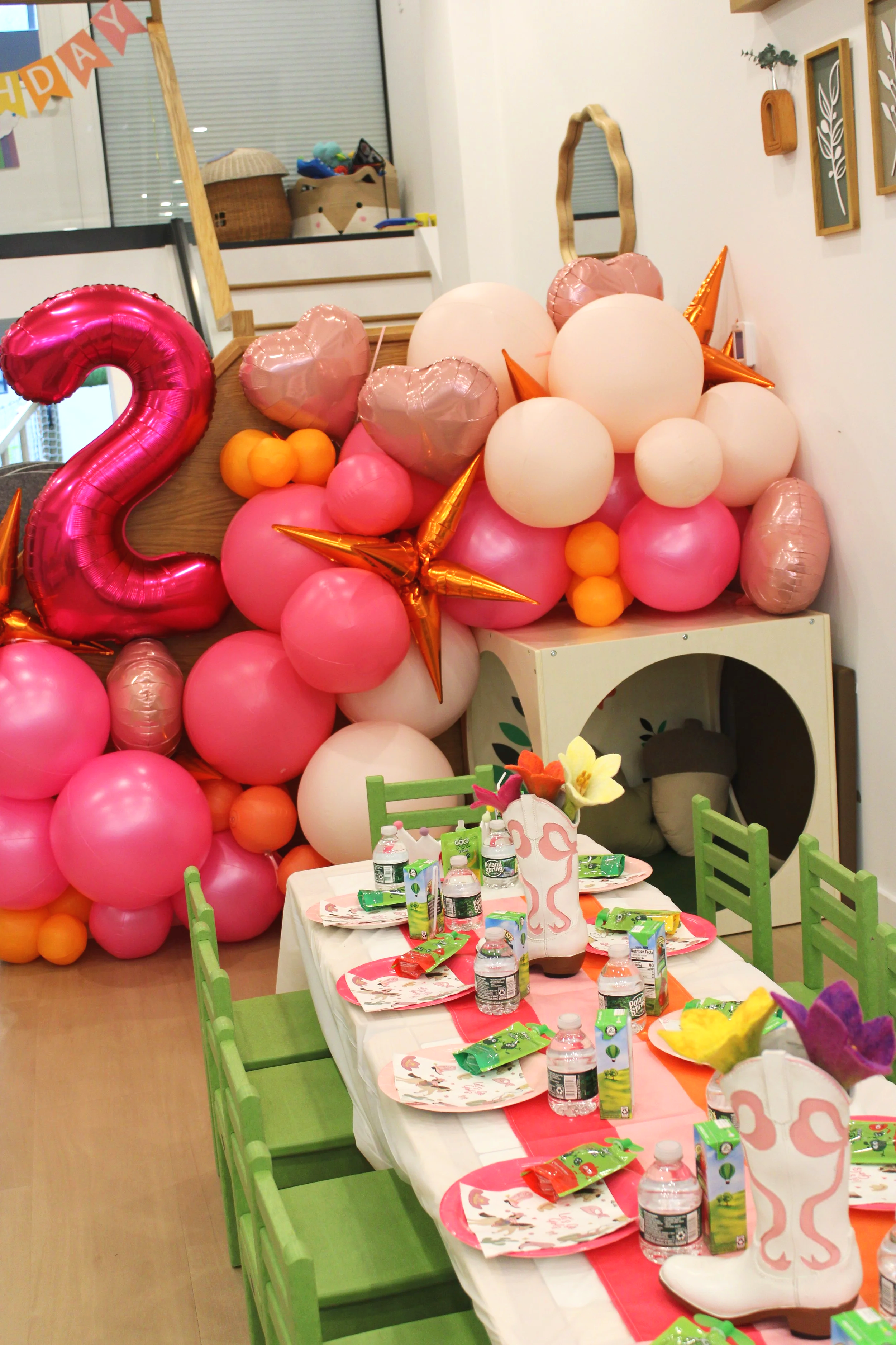 Party decorations with pink, white, and orange balloons, including a large pink number 2 balloon and star-shaped balloons, above a table set for a celebration with plates, juice boxes, water bottles, and table decorations, in a colorful room.