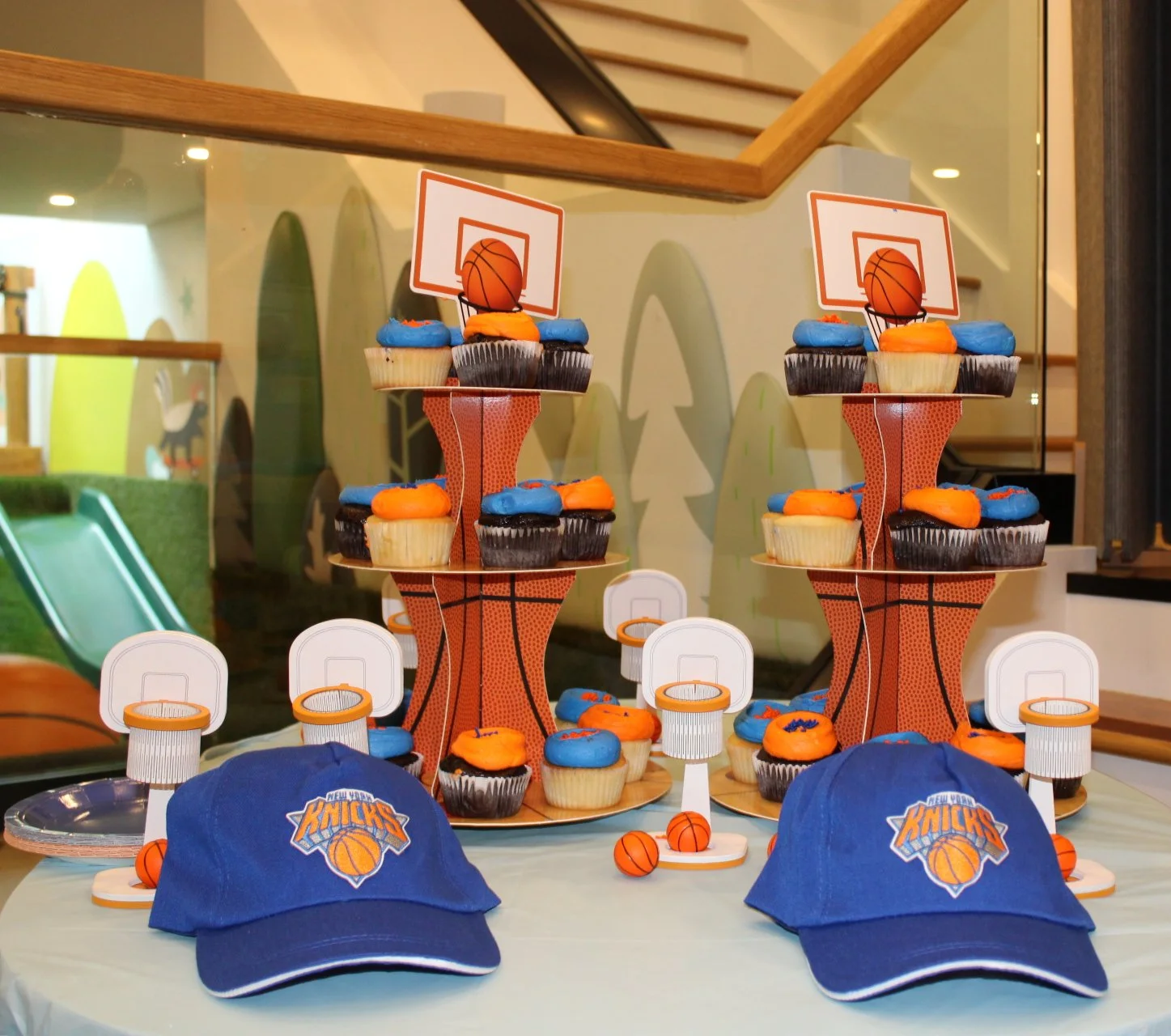 Basketball themed cupcakes on a table with New York Knicks hats and mini basketballs, set in a room with colorful wall art and a staircase in the background.