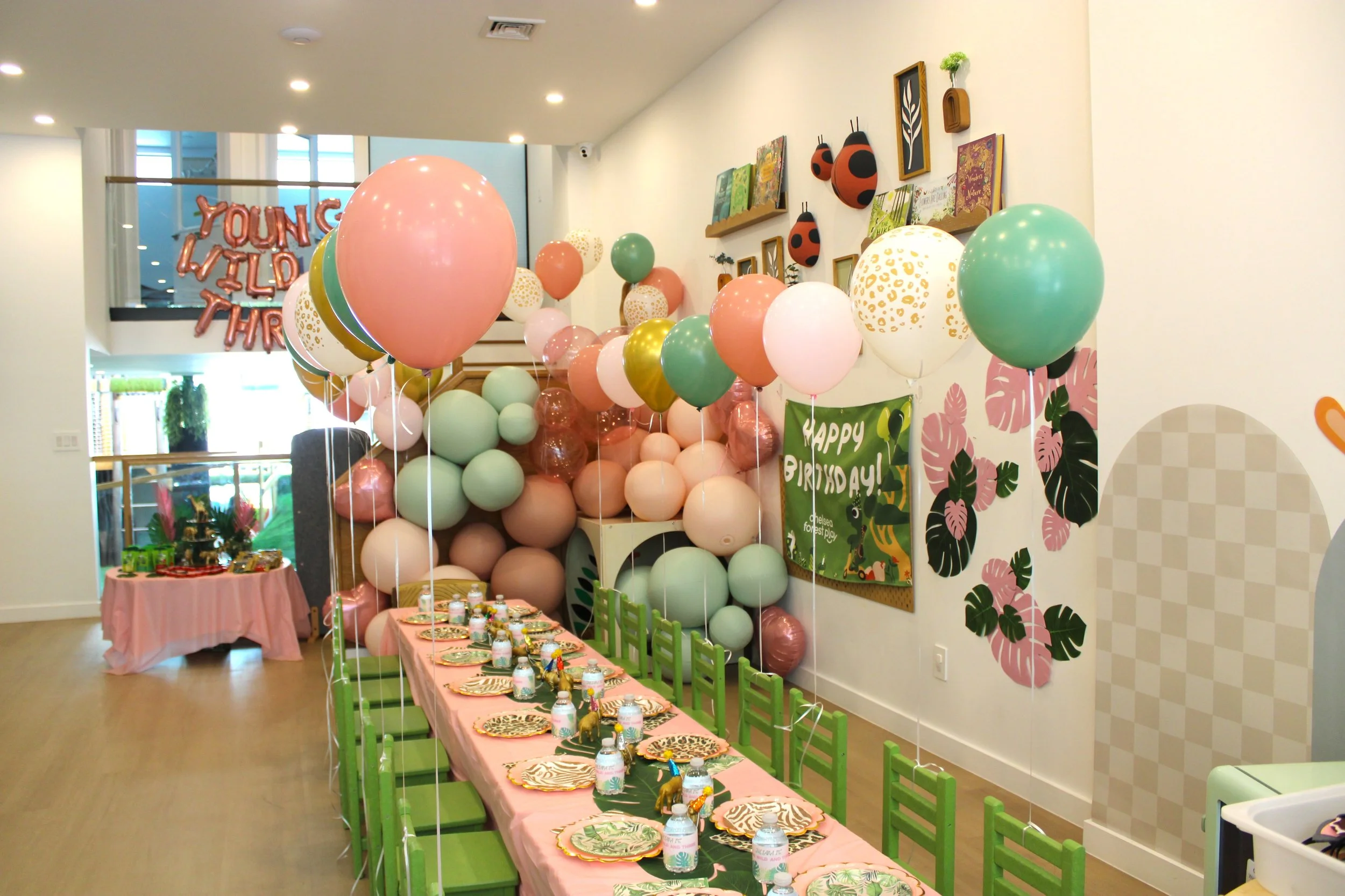 Decorated party room with pink, green, and white balloons, a long table with pink tablecloth, party hats, and themed tableware arranged for a celebration, with a 'Young Wild Three' balloon sign visible in the background.