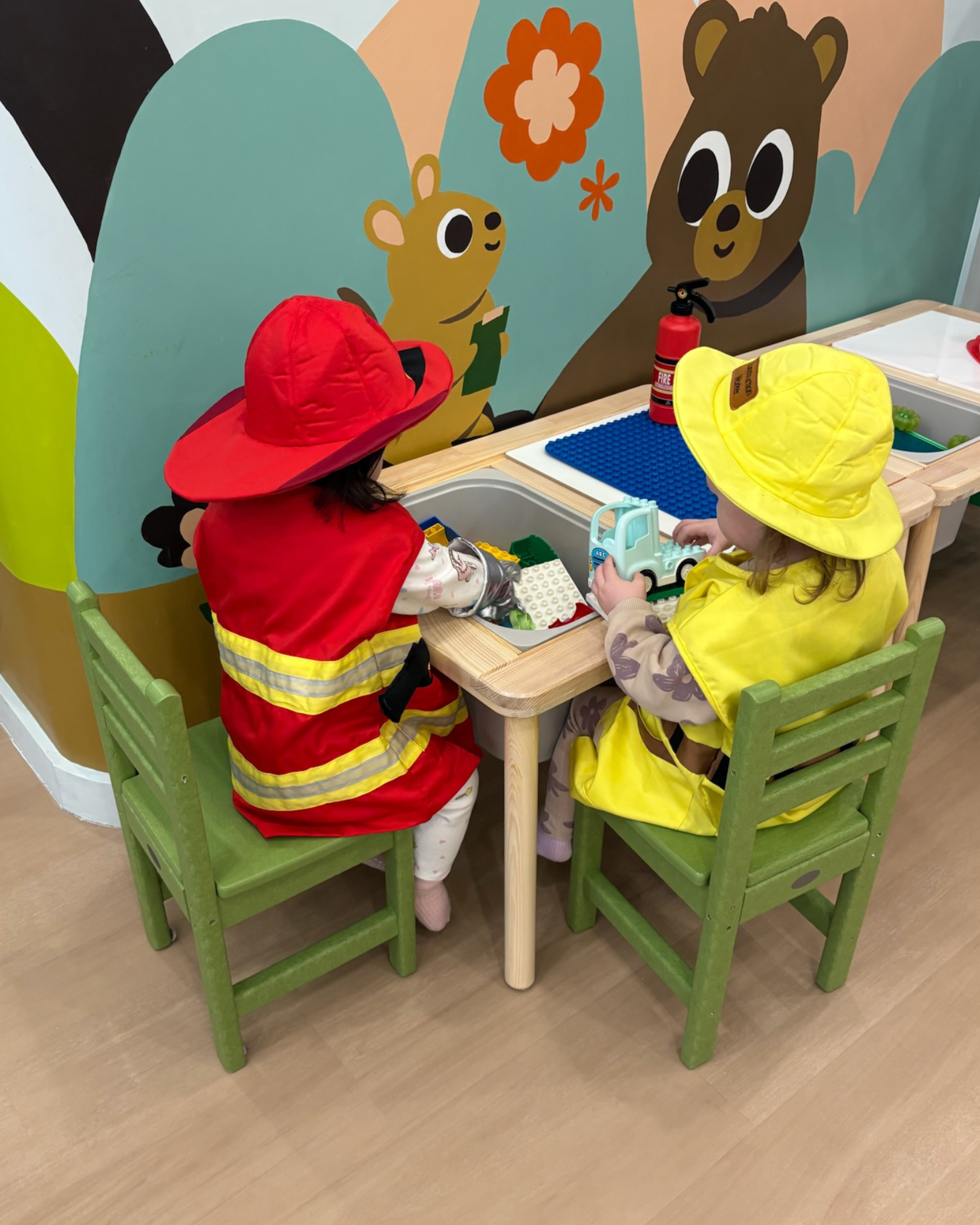 Two children dressed as firefighters playing with toy vehicles at a small table in a colorful playroom. One child wears a red firefighter costume, and the other wears a yellow firefighter costume, both with matching helmets.