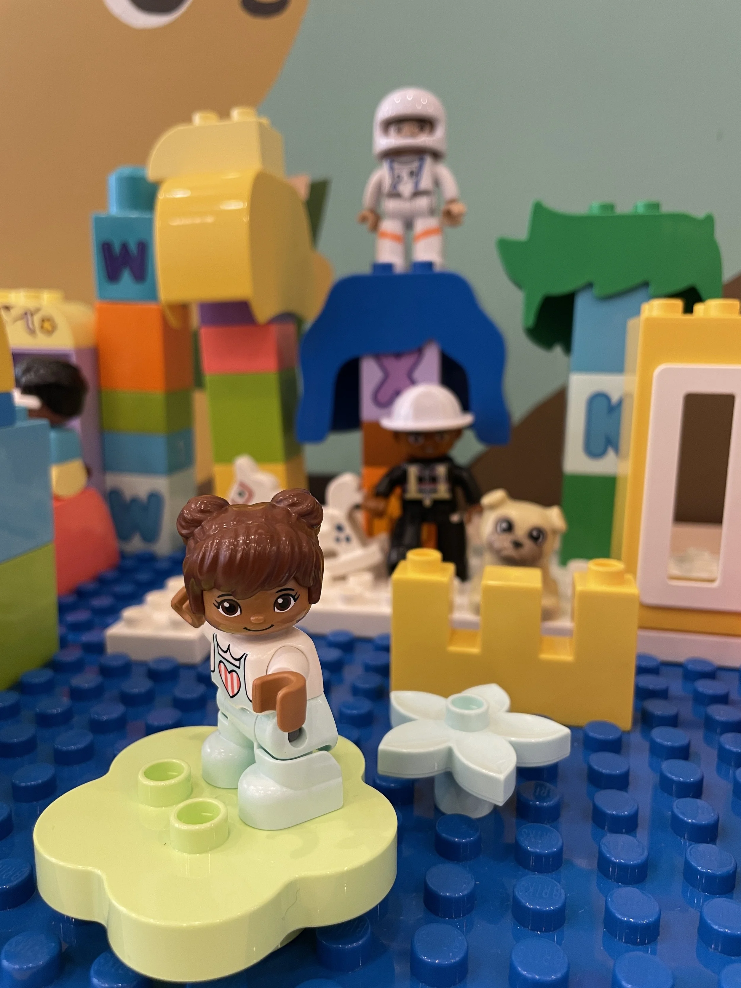 A close-up of Lego figurines and Lego construction elements on a blue baseplate. Front and center is a Lego girl with brown hair, wearing a white dress with a red heart. Behind her is a variety of other Lego figures, including a girl wearing a white helmet and black outfit. In the background, there's a Lego structure with multicolored bricks, including yellow, blue, pink, and green, and a Lego astronaut figure standing on top of a blue arch.
