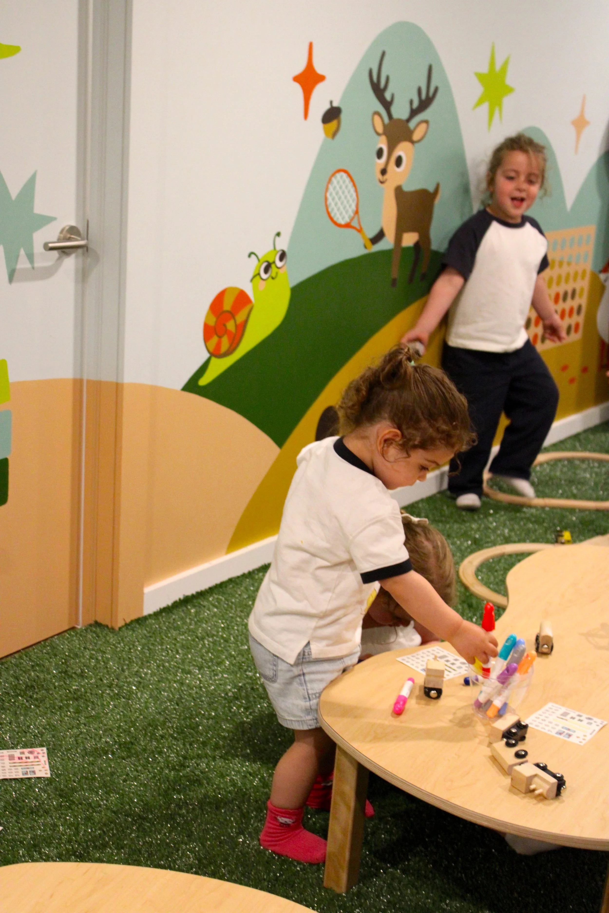 Children playing with educational toys at a table in a colorful classroom with a nature-themed mural on the wall.