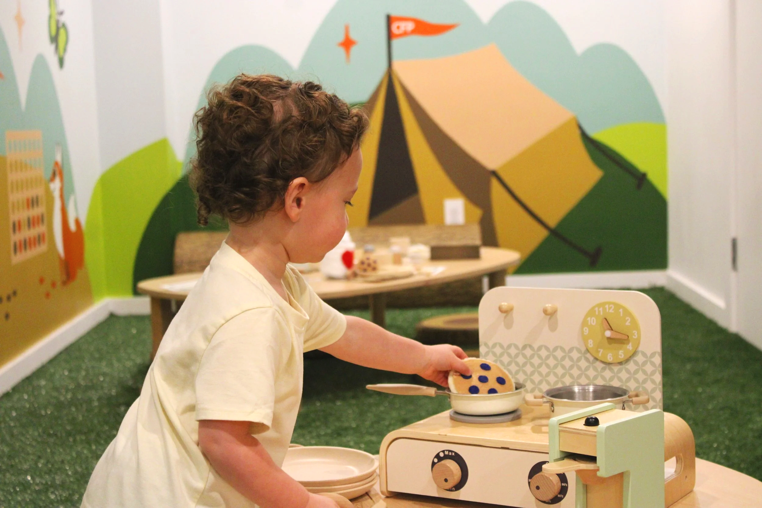 A young child with curly hair in a yellow shirt playing with a wooden toy stove in a colorful room with a mountain and tent mural on the wall.
