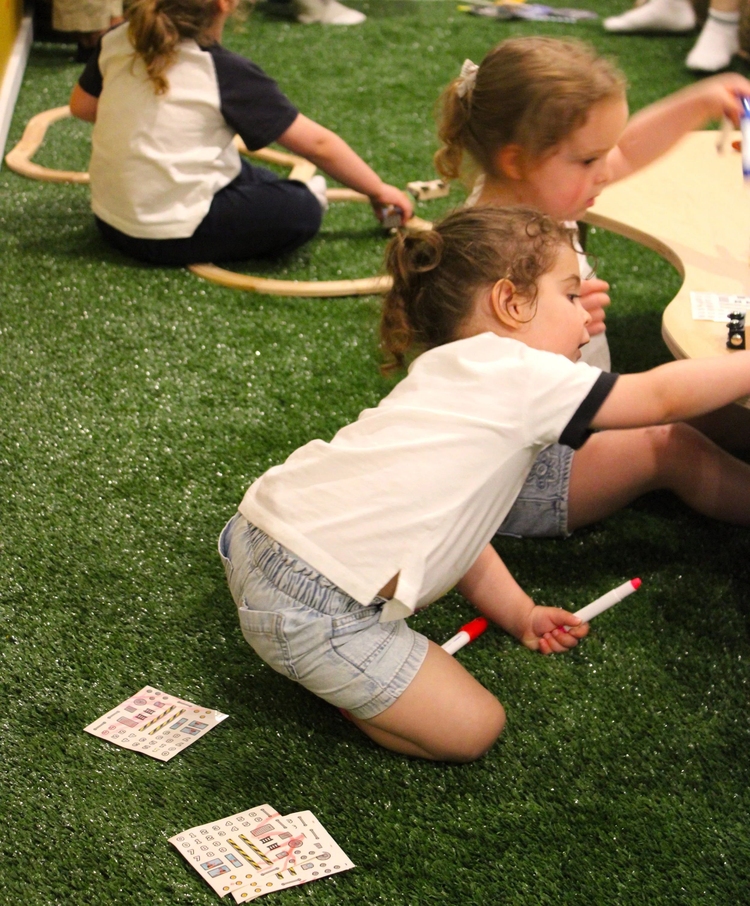 Two young children sitting on a green artificial grass surface, playing with markers and stickers. One girl in a white shirt and shorts is lying on her stomach, holding a red marker, with sticker sheets in front of her. The other girl, with curly hai