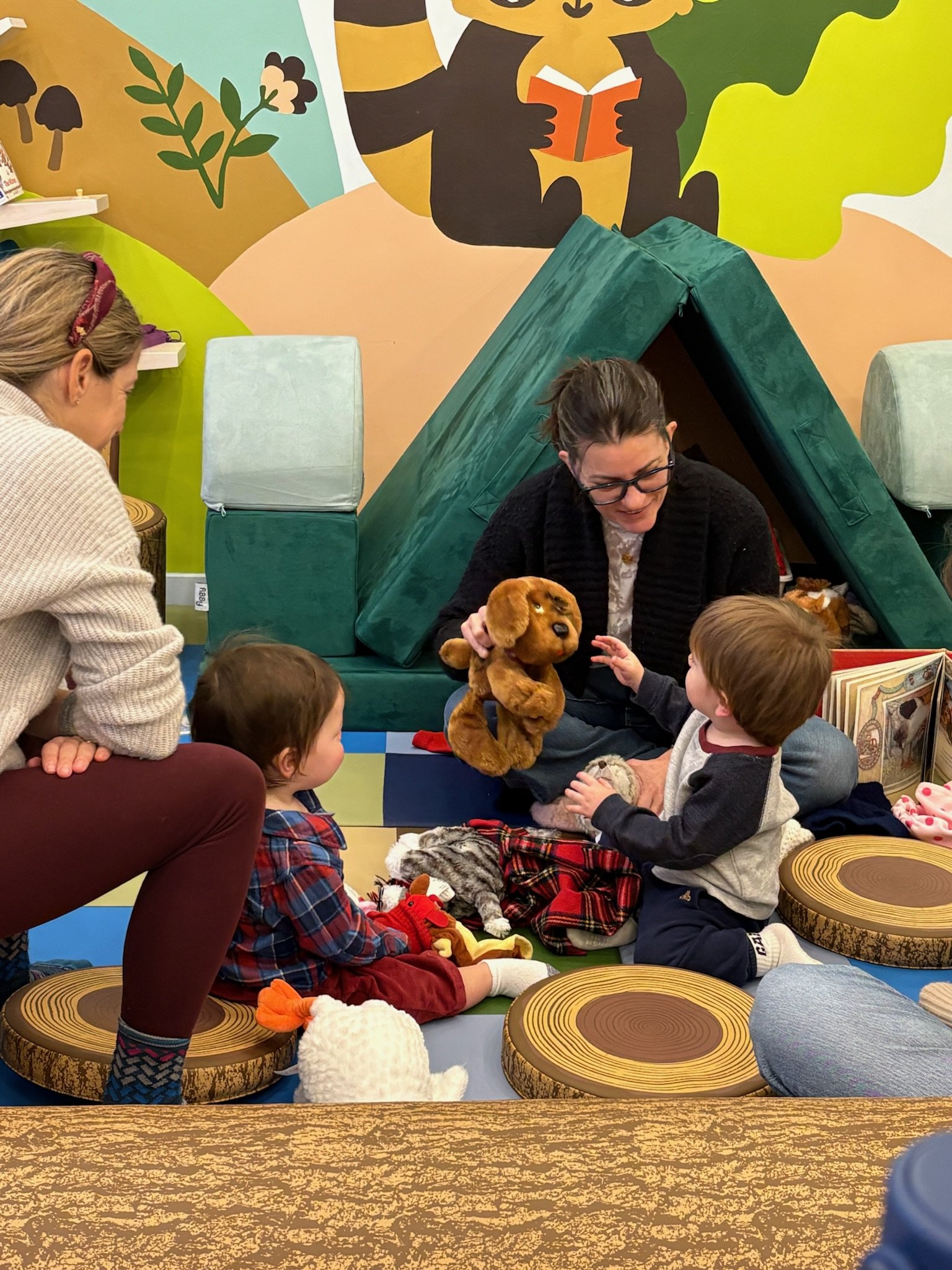 A woman with glasses and dark hair is using a puppet dog to entertain two children sitting on a colorful play mat. One child, with light brown hair and a gray sweater, reaching out to the puppet, while the other, with dark hair, dressed in a plaid sh