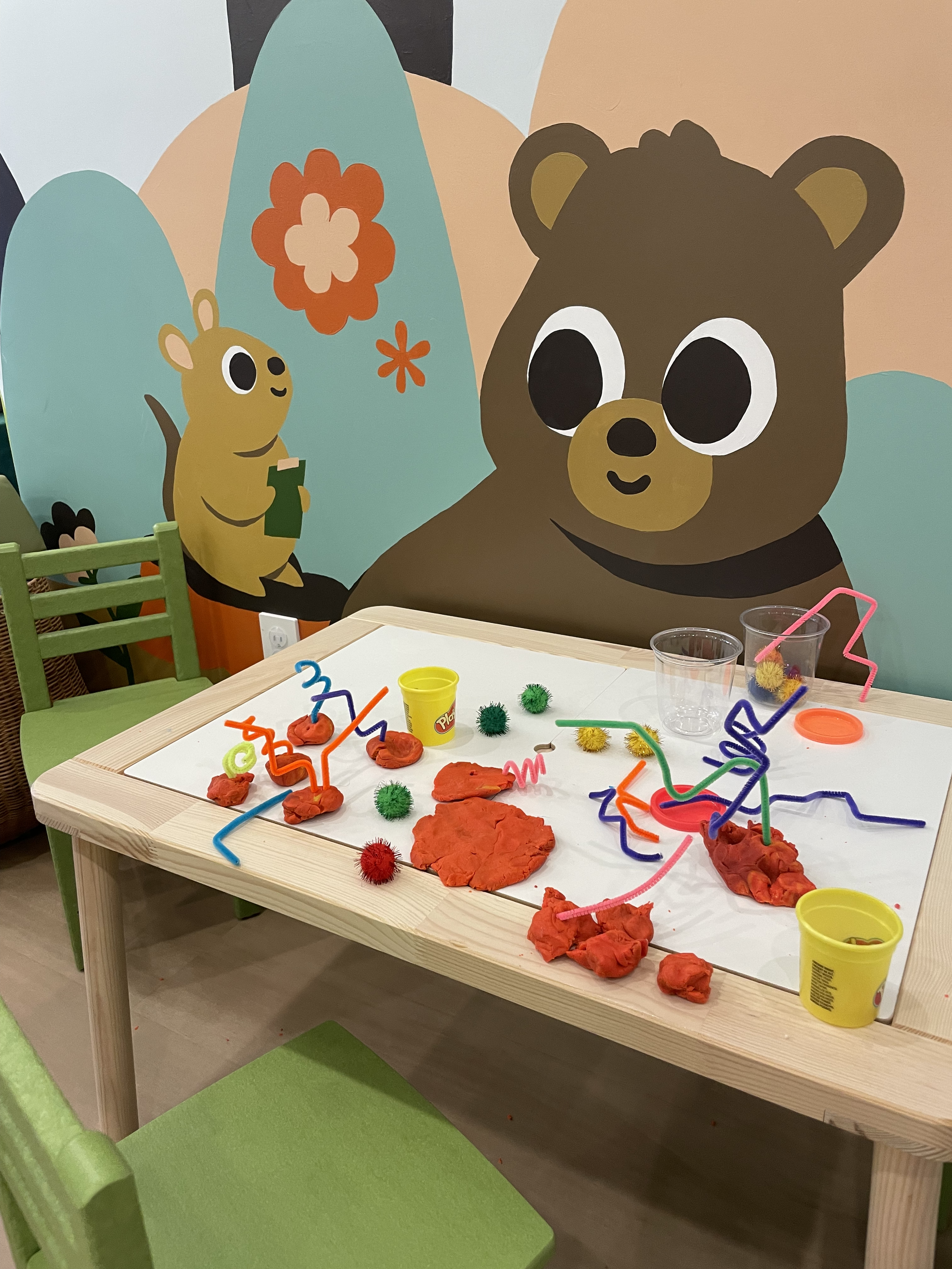 A children's play area with a wooden table covered in red Play-Doh, pipe cleaners, pom-poms, and plastic cups. In the background, there are colorful cartoon animal murals, including a bear and a mouse on a nature-themed wall.