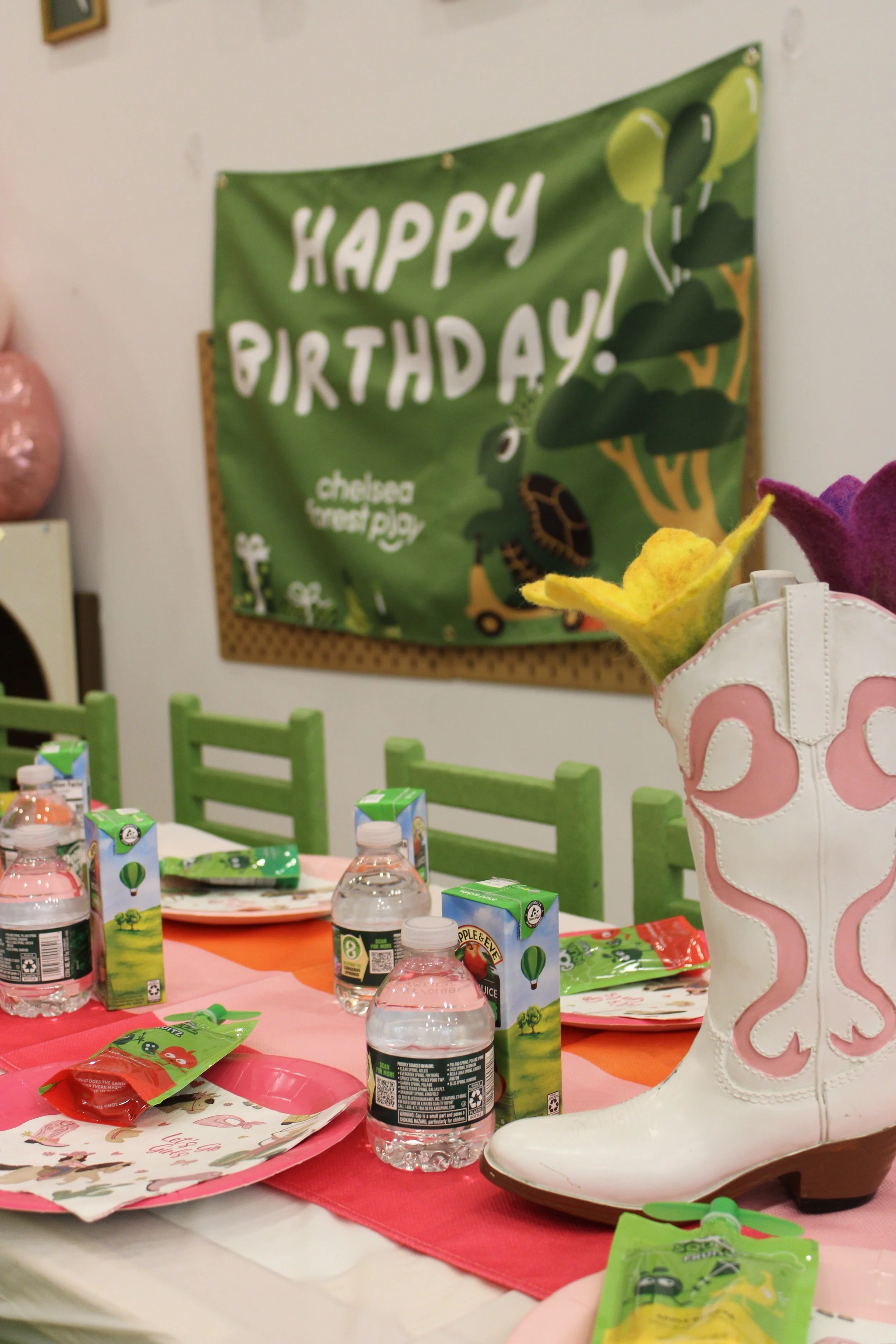 A party table decorated with a pink and orange tablecloth, assorted juice boxes, bottled water, and party favor bags. A green 'Happy Birthday' banner with a dinosaur illustration is on the wall in the background. Pink, purple, and white slippers with a flower design are on the table.