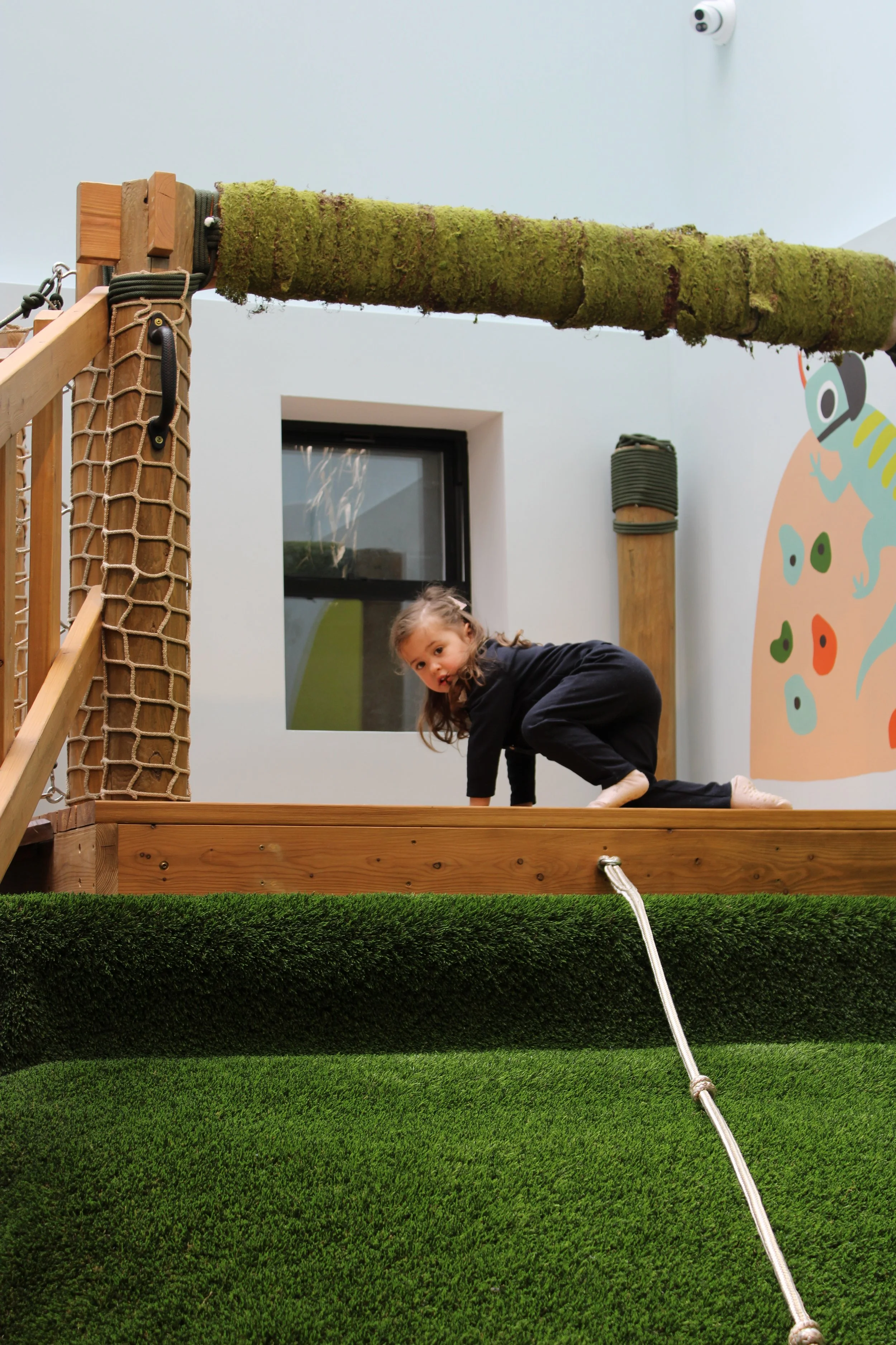 A girl in a dark sweater and black pants is crawling on a wooden platform in an indoor play area. There is a moss-covered horizontal pole overhead, part of the playground structure. The lower part of the image shows artificial green grass, and a white rope is hanging down from the platform. In the background, a colorful wall decoration with a gecko illustration and a window are visible.