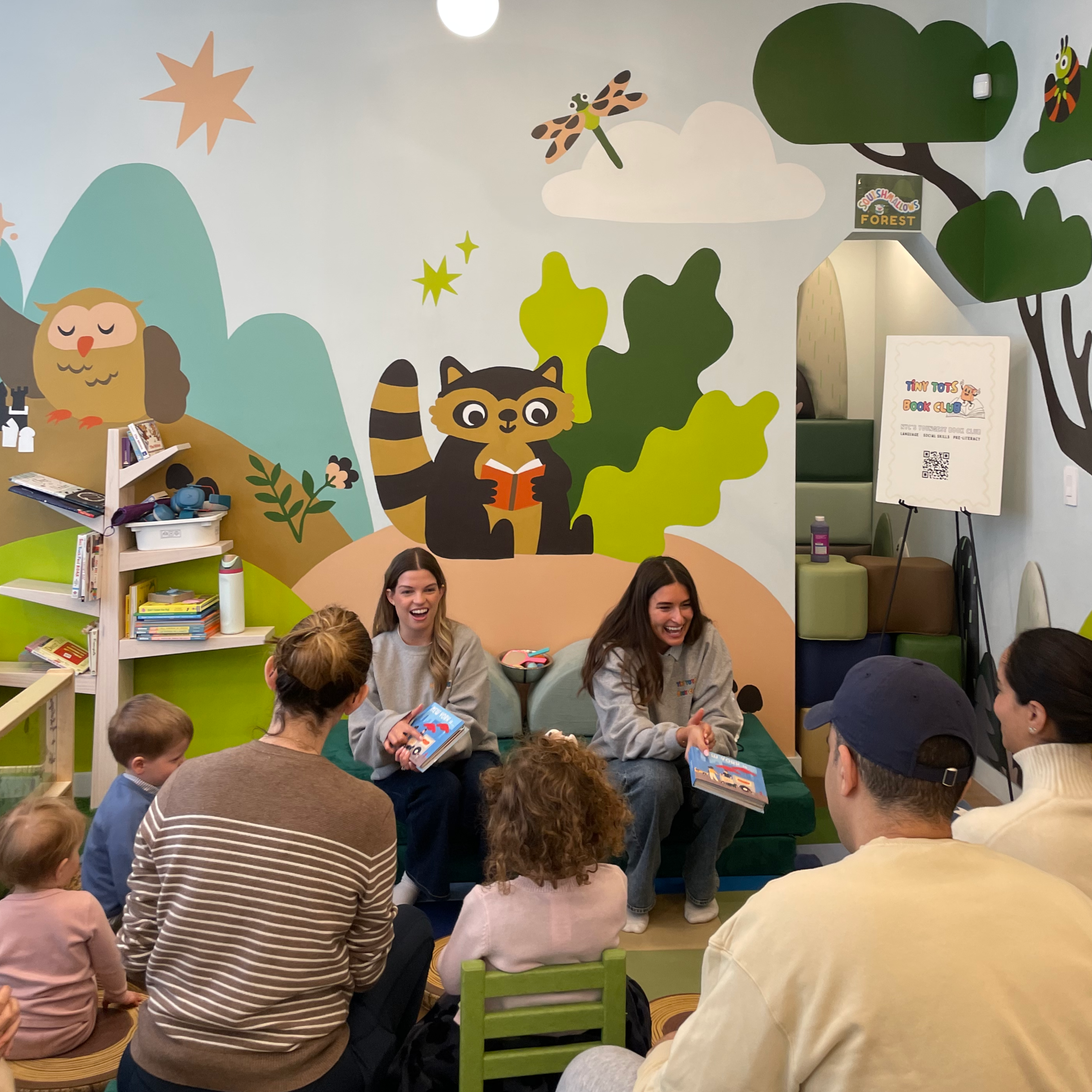 A group of children and adults are seated on the floor of a children's library or storytime room, engaging in a storytime or group activity. The room features a colorful mural with cartoon animals, trees, clouds, and stars on the wall. There are bookshelves filled with books on the left side and a poster on an easel at the back of the room.