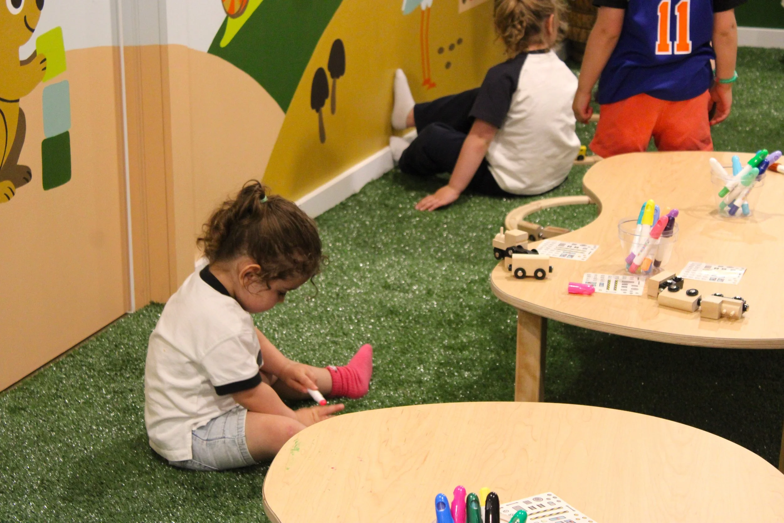 Children playing and drawing in an indoor play area with colorful wall art, small wooden trains, and a round table with markers