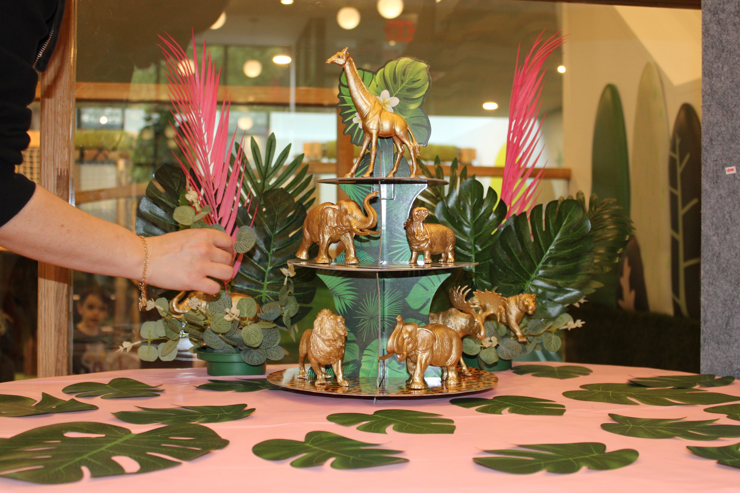 Gold animal figurines of a giraffe, elephant, lion, and panther are arranged on a tiered display surrounded by tropical leaves and pink decorative plants.