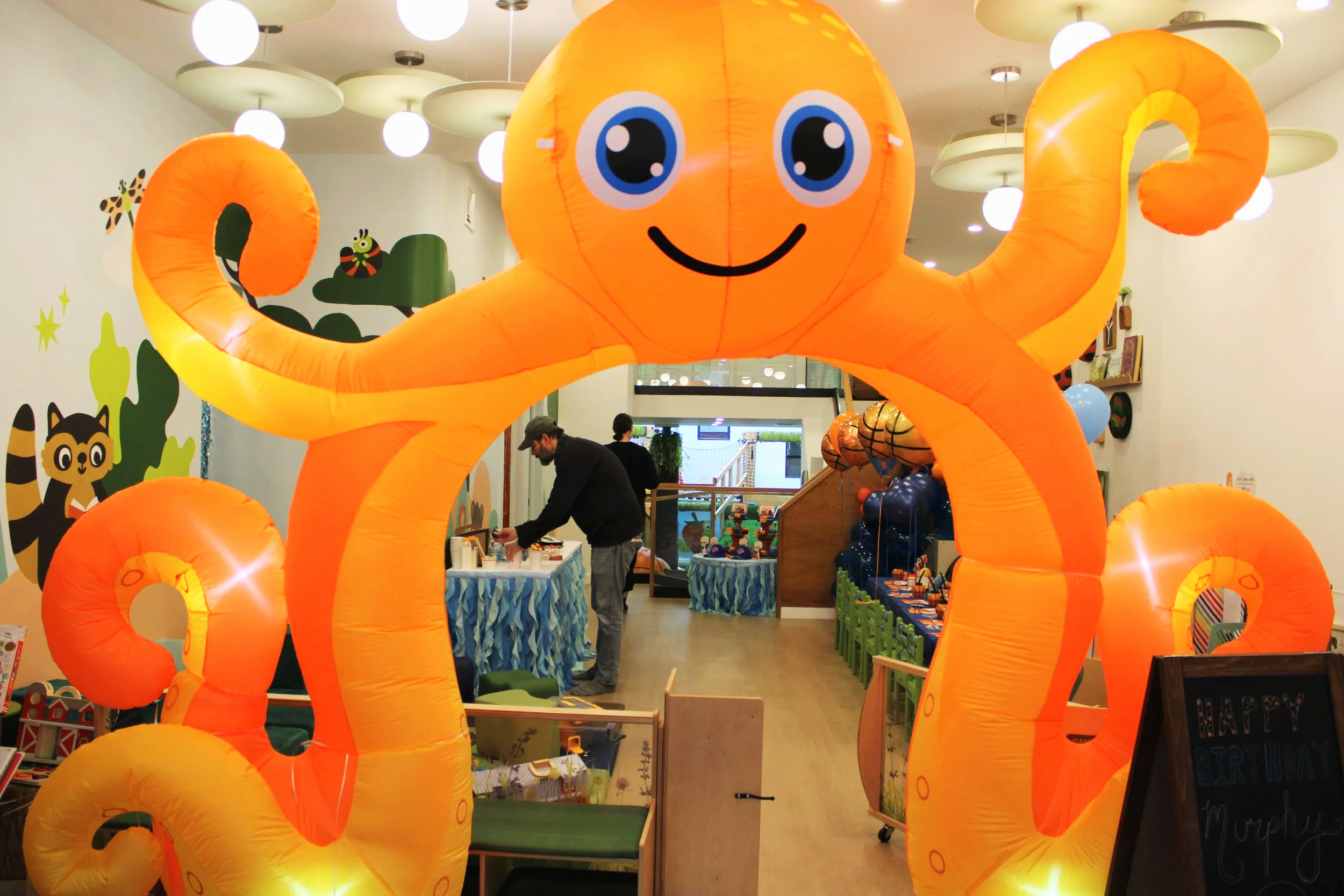 A large inflatable octopus with a smiling face and big blue eyes, orange in color, forming an entrance arch at a children's event or party indoors.