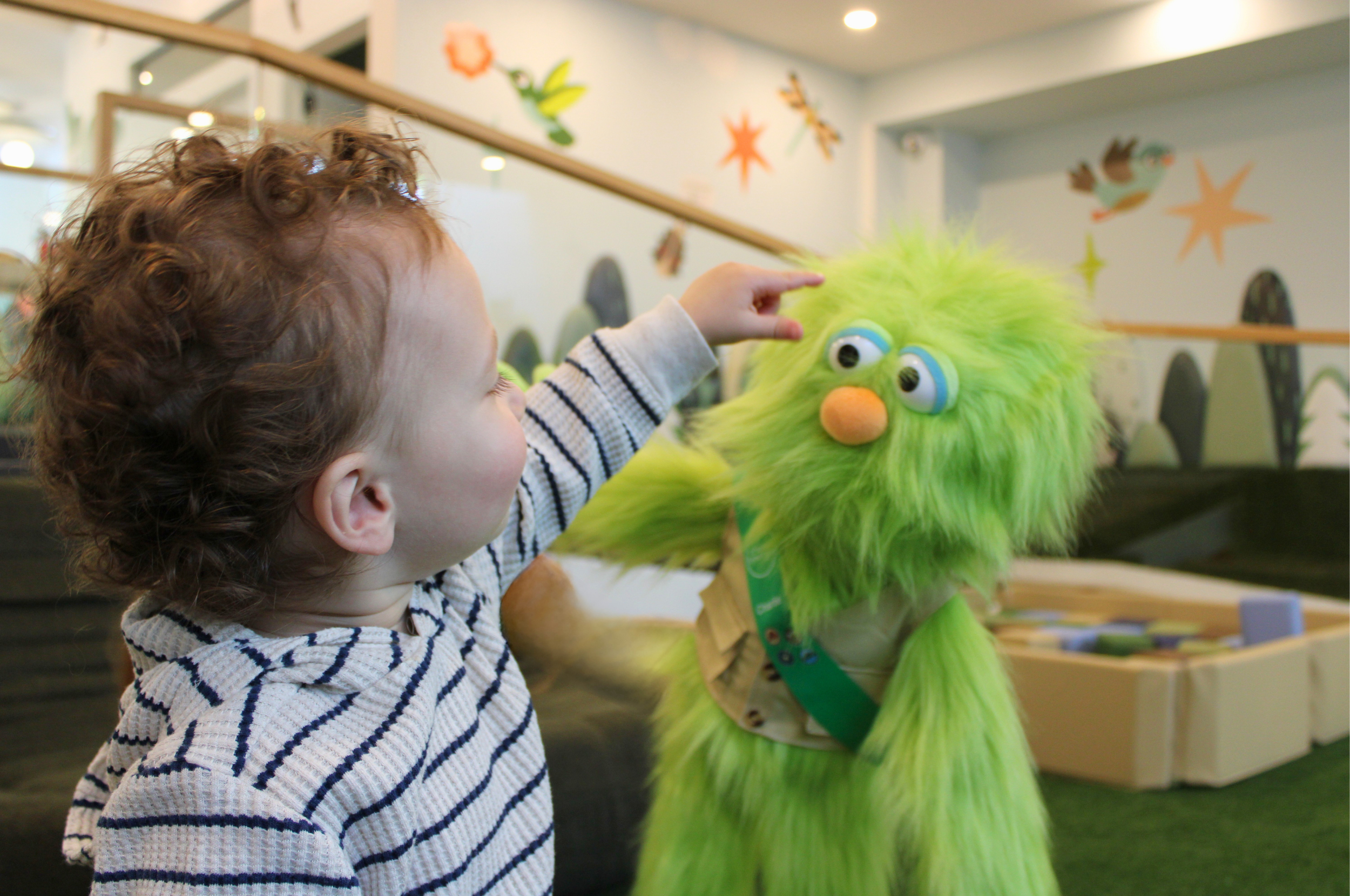 A young child with curly hair points to a green furry puppet with big googly eyes and an orange nose, in an indoor setting with colorful wall decorations.