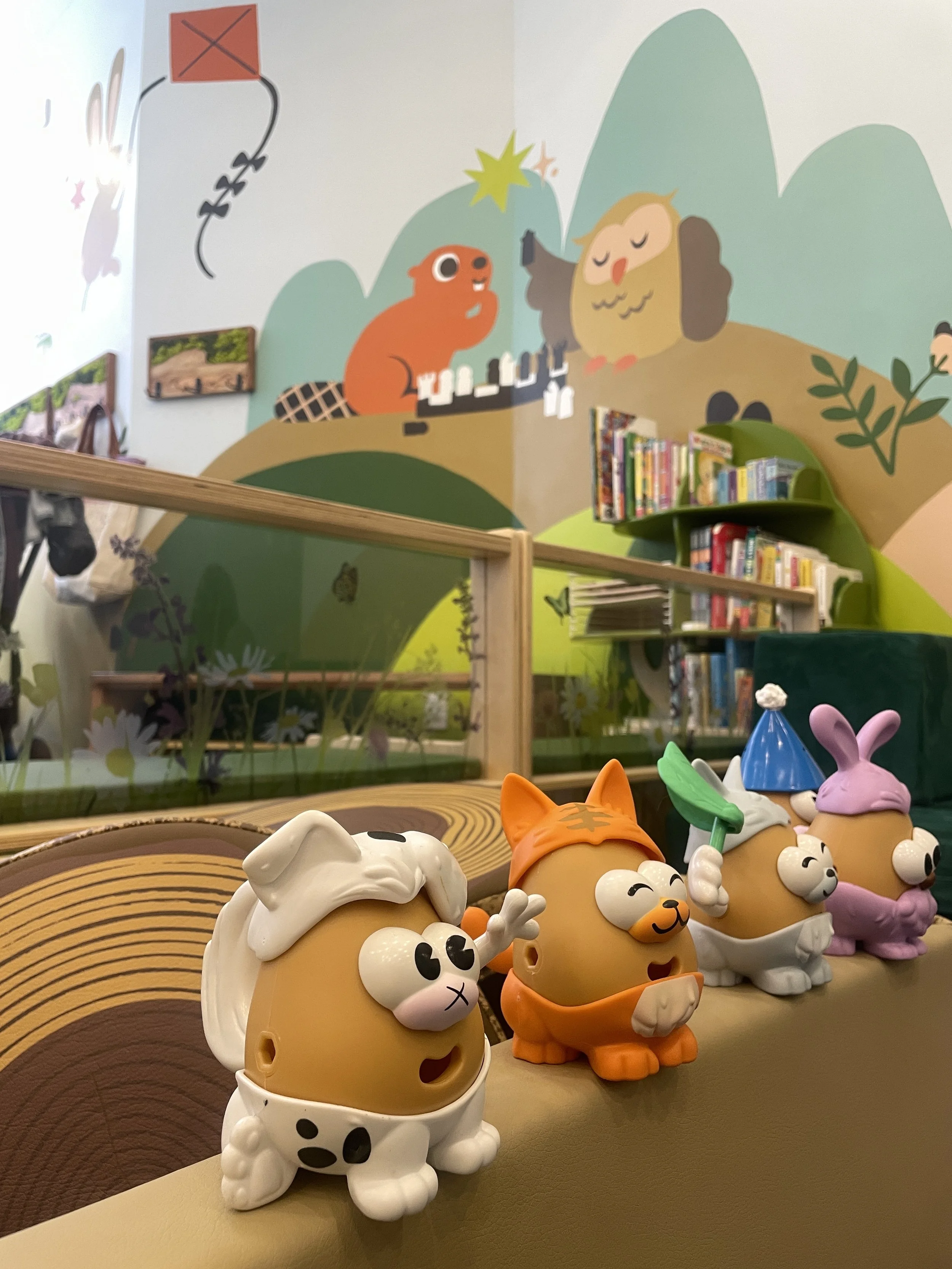 Colorful animal figurines with cartoonish features on a table, including a white dog, an orange cat, a white cat with a green leaf hat, and a purple bunny with a pink hat, against a background with books and a mural of a squirrel and an owl.