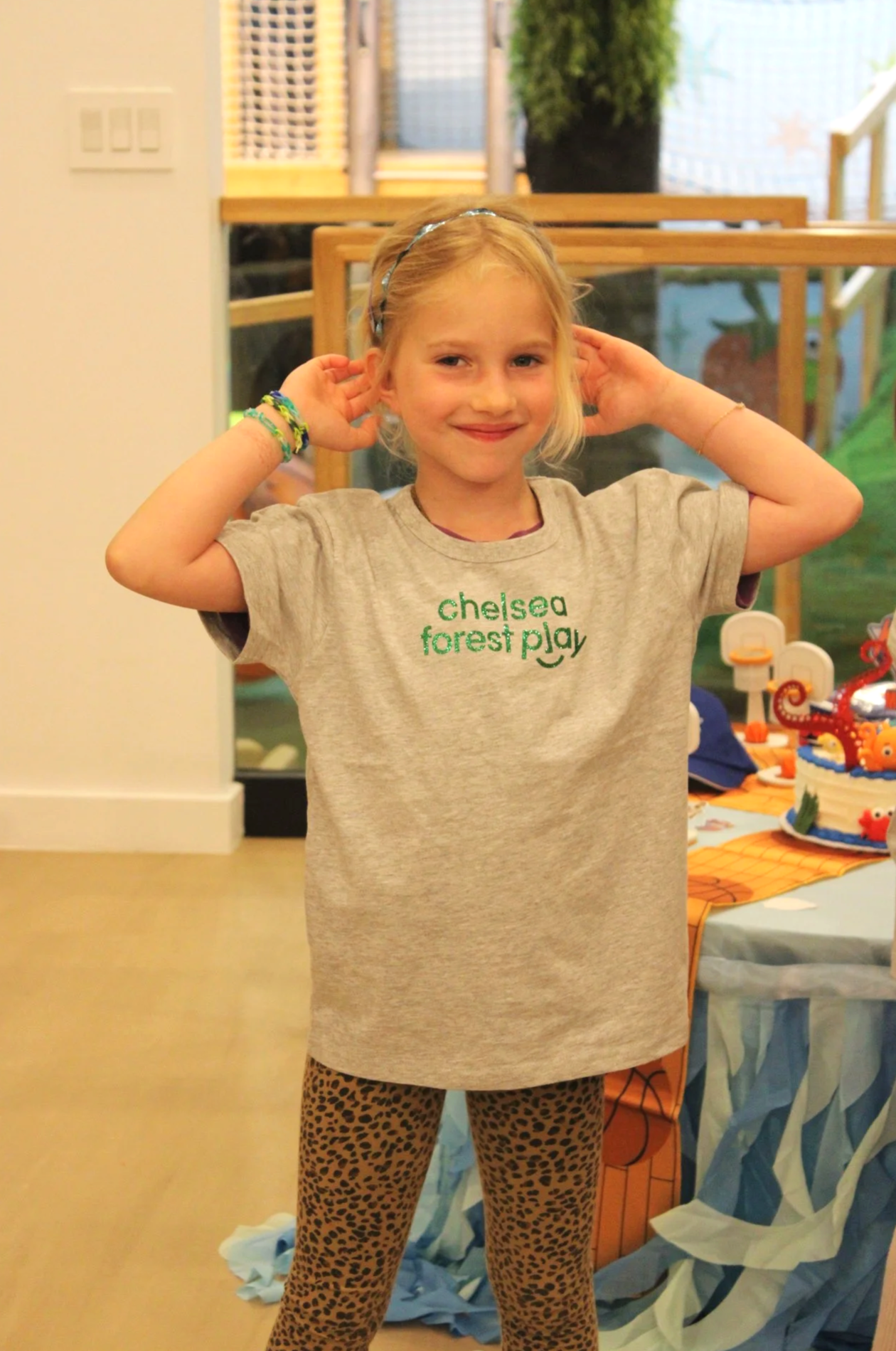 A young girl with blonde hair, wearing a gray t-shirt with green text and leopard print pants, posing with her hands near her ears at a birthday party. There are decorations, a cake, and a table in the background.