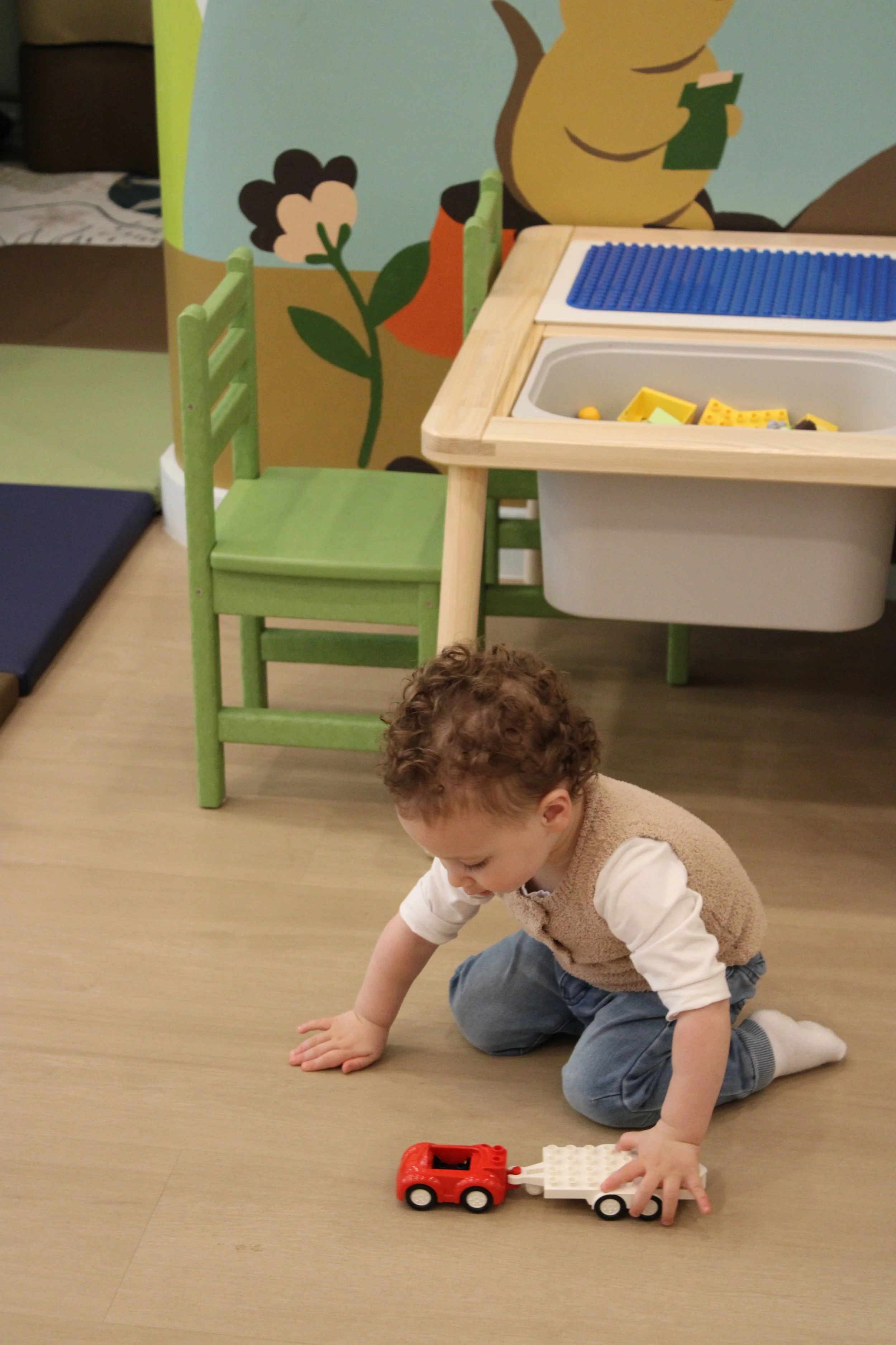 A young boy crawling on the floor playing with a small toy truck. There is a green chair, a wooden table with building blocks, and a colorful mural featuring cartoon characters in the background.