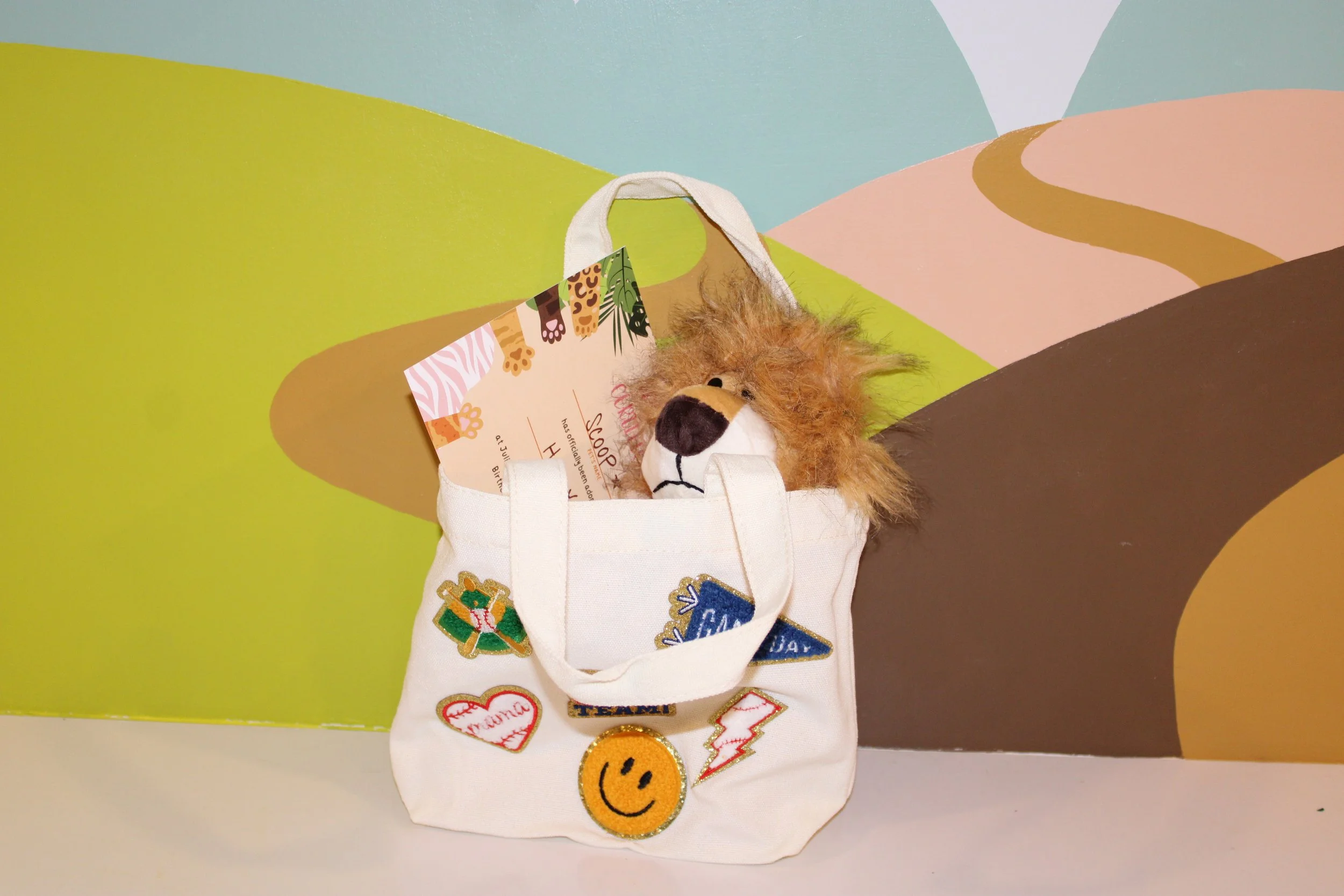 A white tote bag decorated with various colorful patches, including a heart, smiley face, lightning bolt, and other designs, hanging in front of a vibrant, abstract landscape painted on the wall.