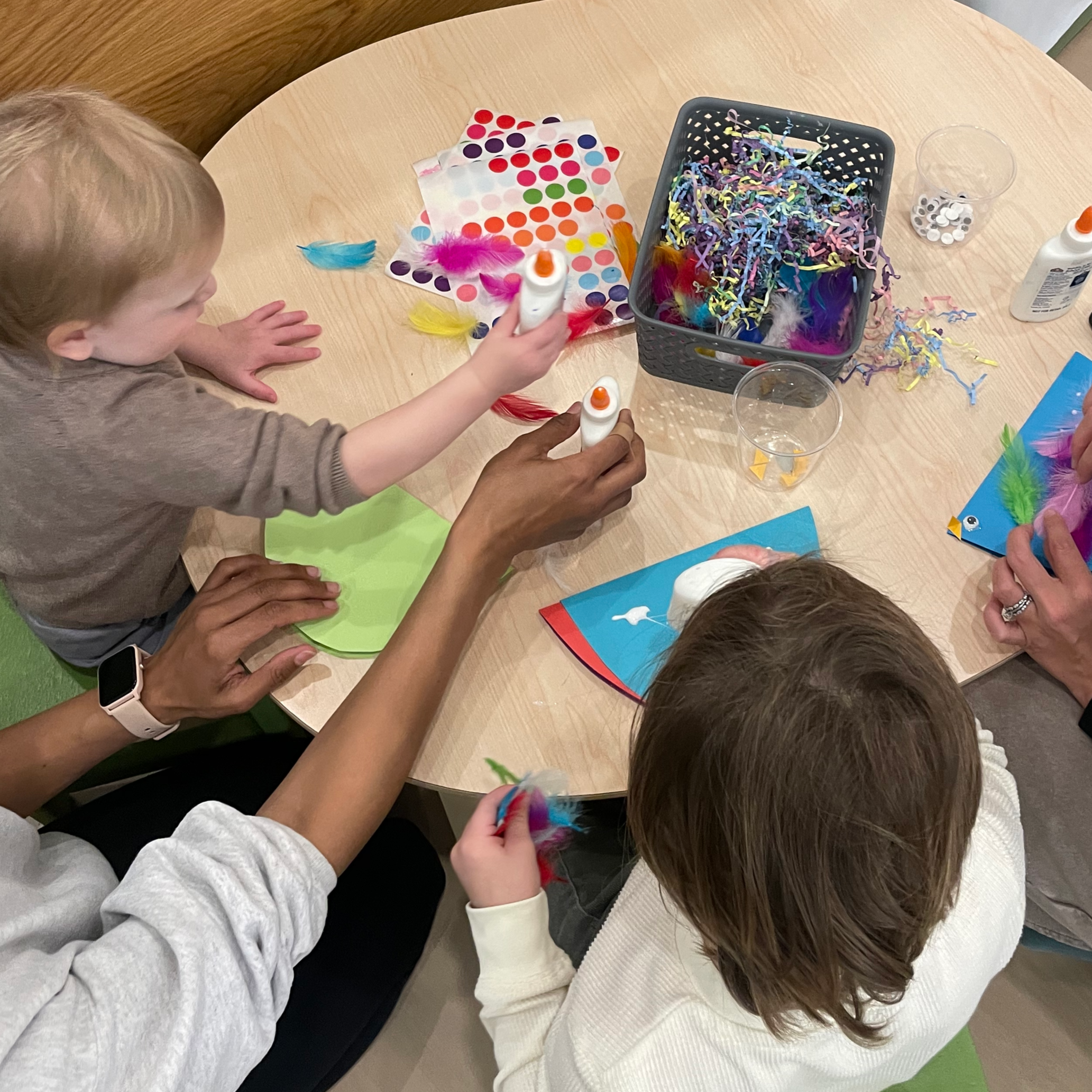 Kids and adults at a table making arts and crafts, with colorful feathers, paper, glue, and a paper craft of a bird.